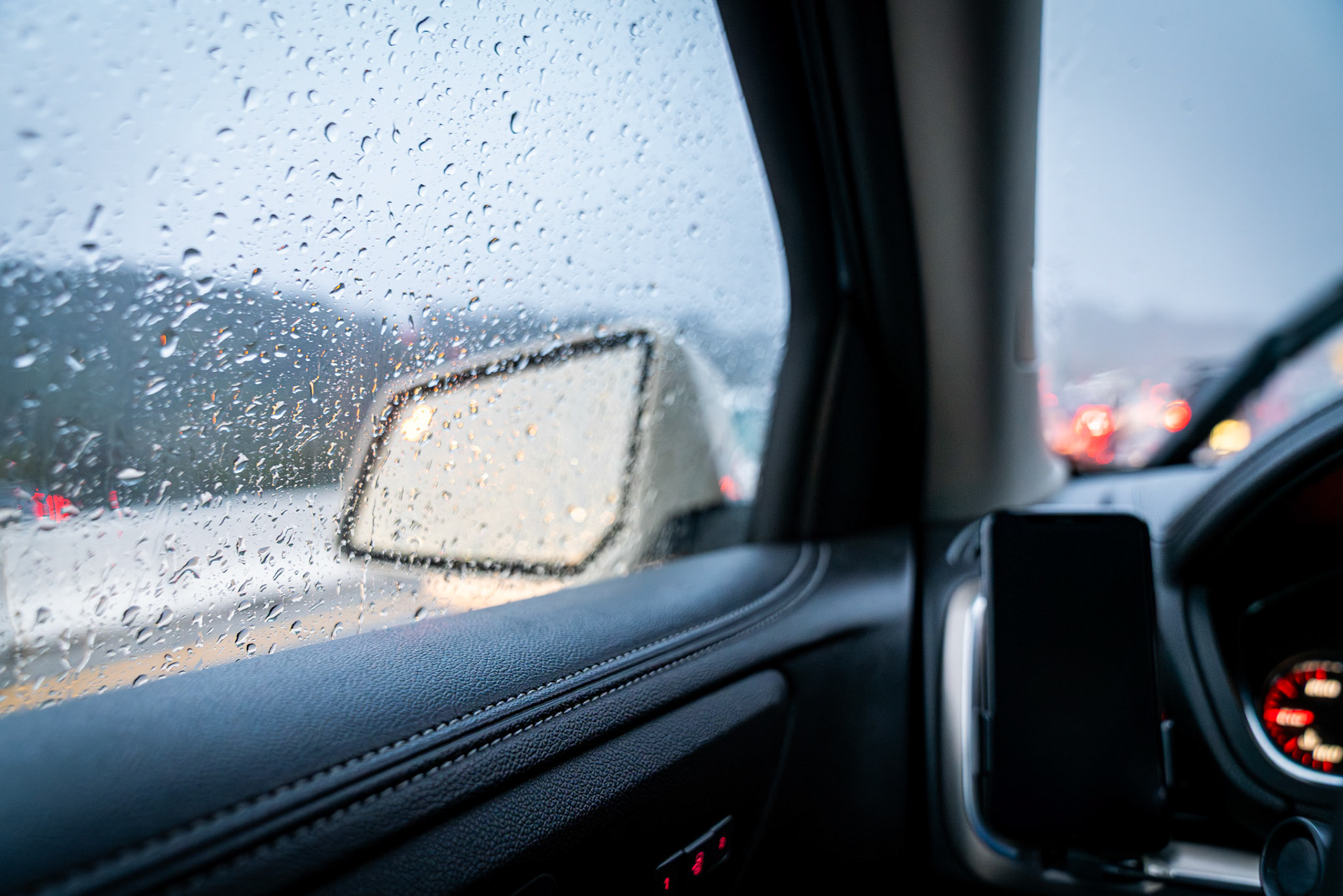 View out a car window through rain drops that blur the freeway and traffic outside the car. A phone is visble mounted on the car vent.
