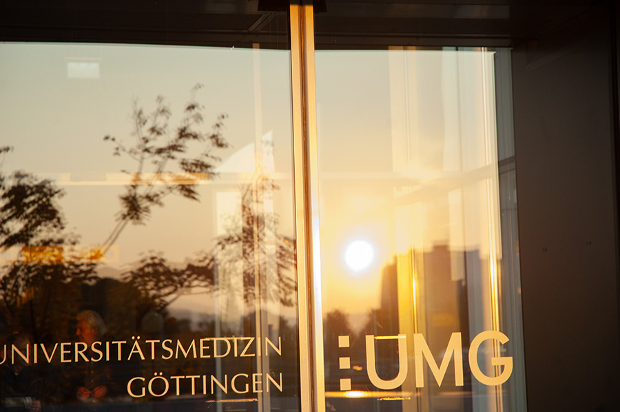 eingangstür mit der aufschrift universitätsmedizin göttingen. im glas spiegelt sich der sonnenuntergang sowie bäume
