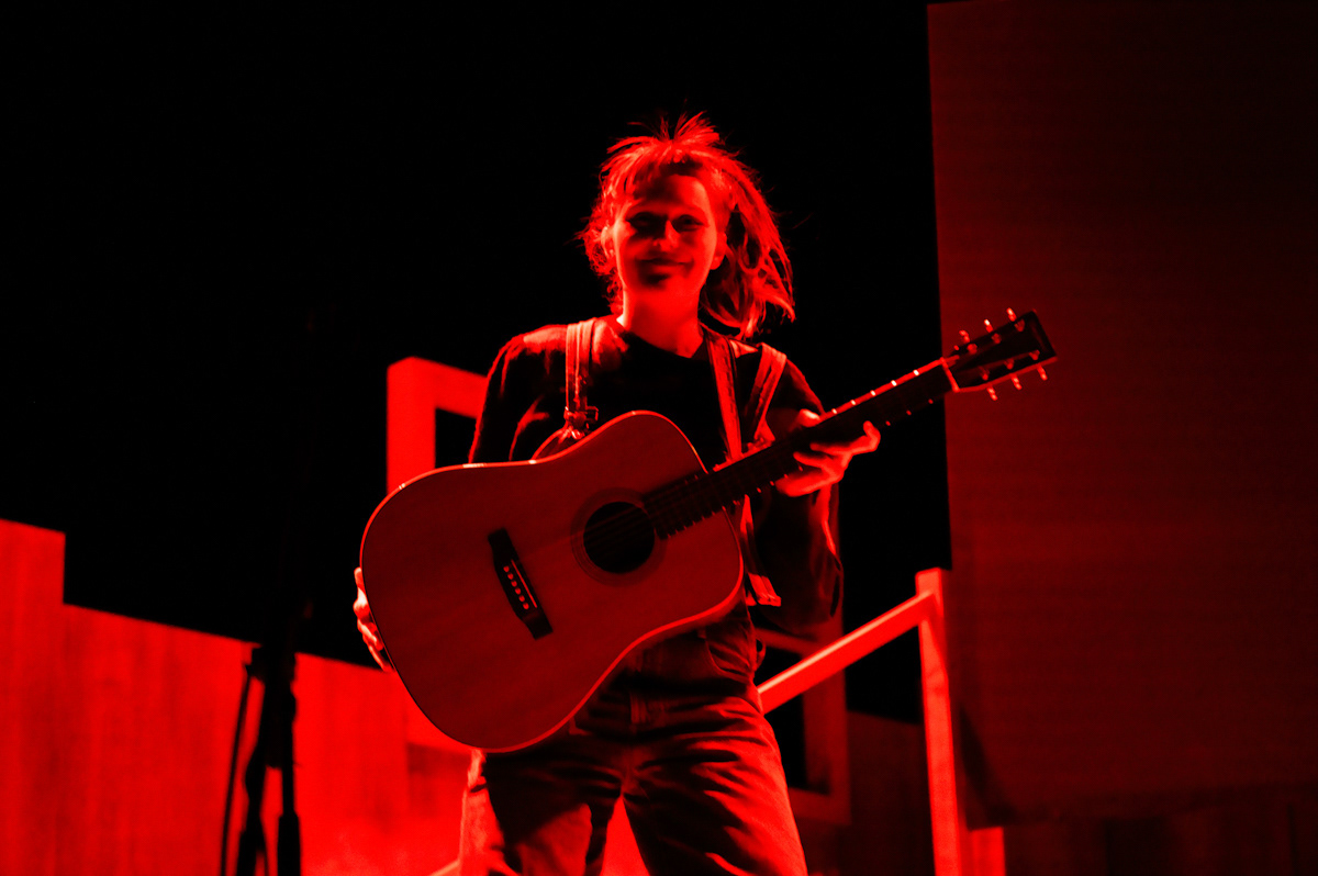 woman on stage with a lot of energy. smiling and with a guitar. red light