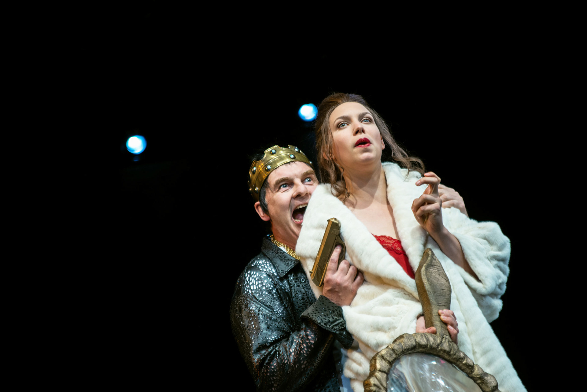 junges schauspielhaus , a man with a weapon standing behind a woman and he is jelling. its an theater scene. ein mann mit einer krone und einer waffe in der hand steht schreiend hinter einer frau, die eine gr0ße lupe in der hand trägt. es ist eine theaterszene