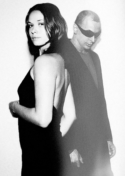analoges foto. eine frau sieht halb mit dem rücken zum betrachter, und schaut lächelnd in die kamera. hinter ihr steht ein mann mit sonnenbrille leicht verdeckt, und sieht auf den boden