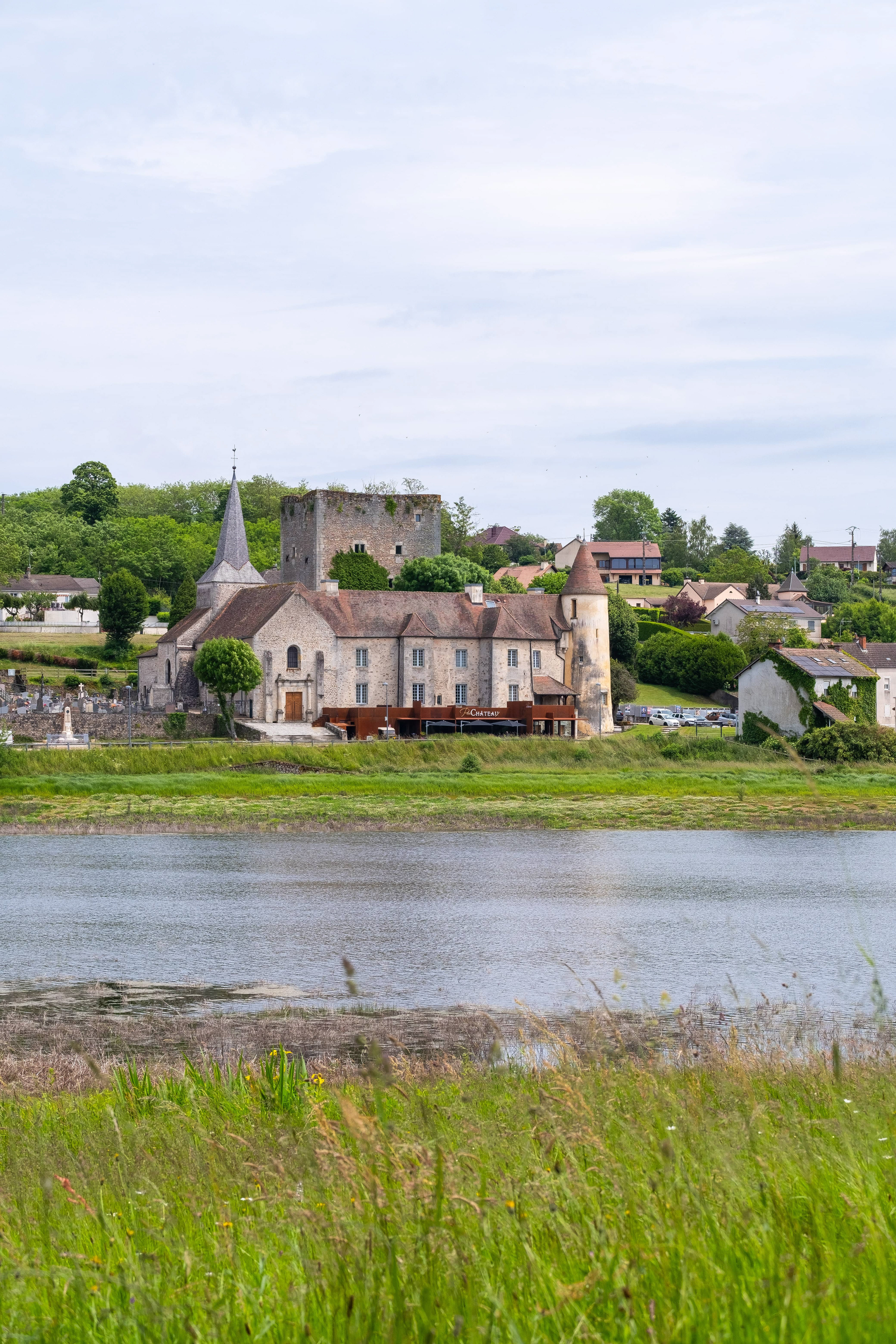 Vue du Restaurant du Château en Saône-et-Loire, Bourgogne, situé au bord d'un lac avec une architecture historique en pierre et une tour, entouré de verdure et d'un paysage pittoresque de village.