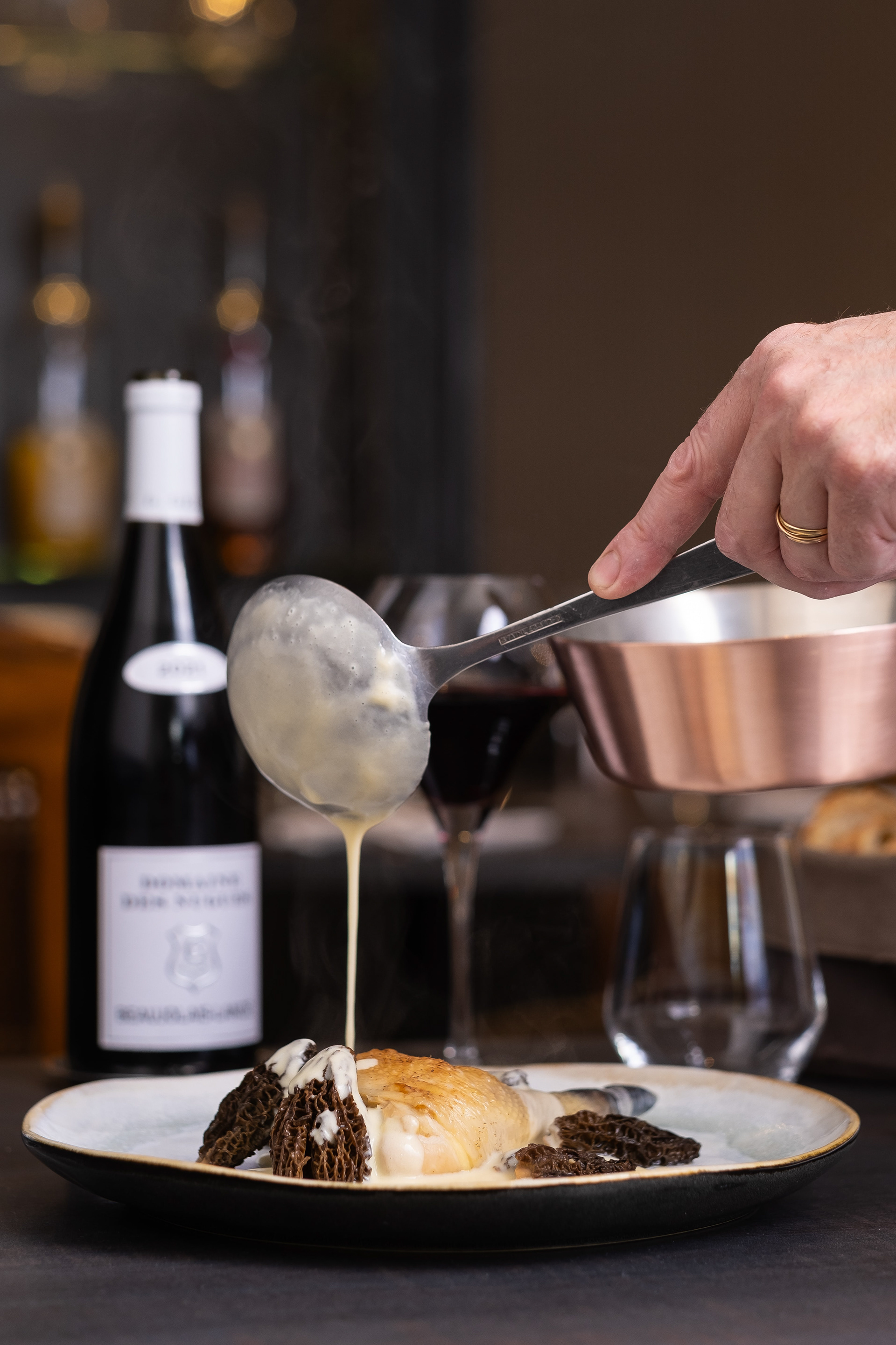 Main d'un chef versant une sauce crémeuse sur une cuisse de volaille de bresse accompagné de morilles, dans une assiette en céramique élégante. En arrière-plan, une bouteille de vin blanc et un verre de vin rouge sont visibles, ajoutant une touche de sophistication à la scène. Le geste délicat du chef, la texture de la sauce et les détails des morilles créent une ambiance gastronomique raffinée