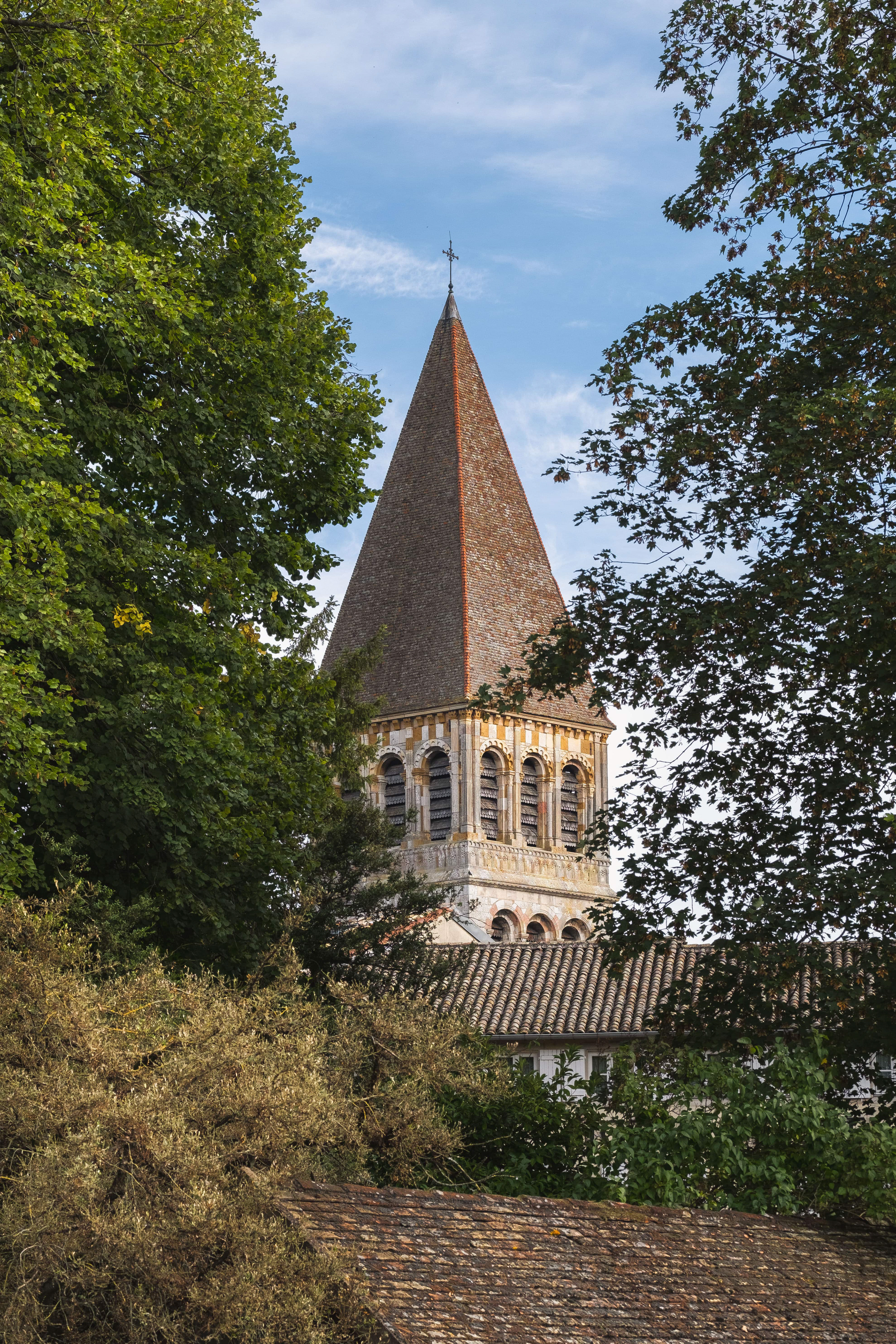 Détails architecturaux du clocher de l'Abbaye de Tournus entouré de verdure luxuriante, avec arcs en pierre et toit en tuiles sous ciel bleu clair. Patrimoine historique et attrait touristique de Saône-et-Loire, Bourgogne. Photographie touristique par Etienne Gatheron.