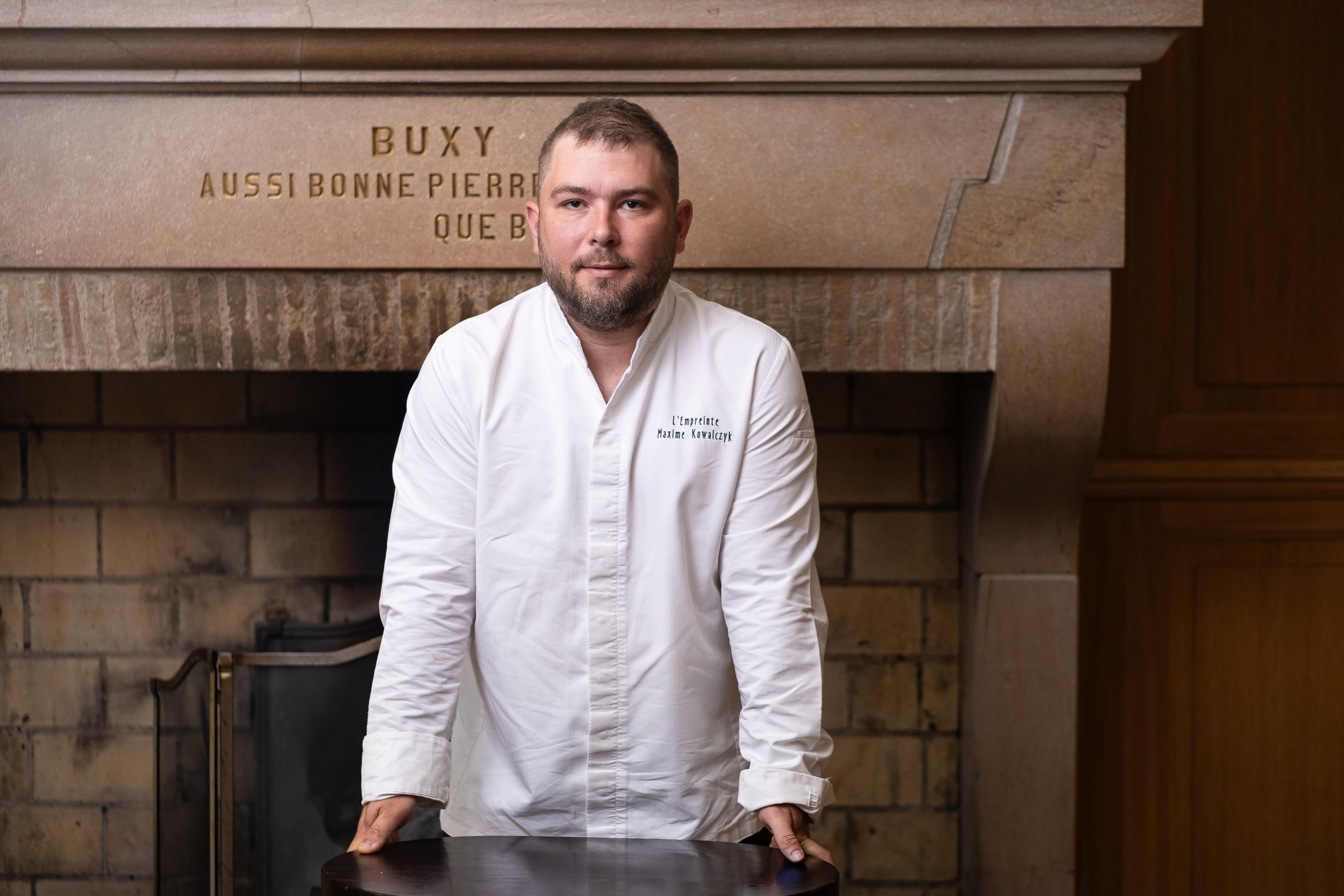 Portrait de Maxime Kowalczyk, chef du restaurant L'Empreinte à Buxy, Saône-et-Loire, Bourgogne, reconnu par Gault et Millau. Debout devant une cheminée en pierre ornée de l'inscription 'Buxy Aussi Bonne Pierre Que Bon Vin', il porte une veste de chef blanche avec le nom de l'établissement brodé. Cette image capture l'élégance et le professionnalisme du chef dans un cadre historique et chaleureux.