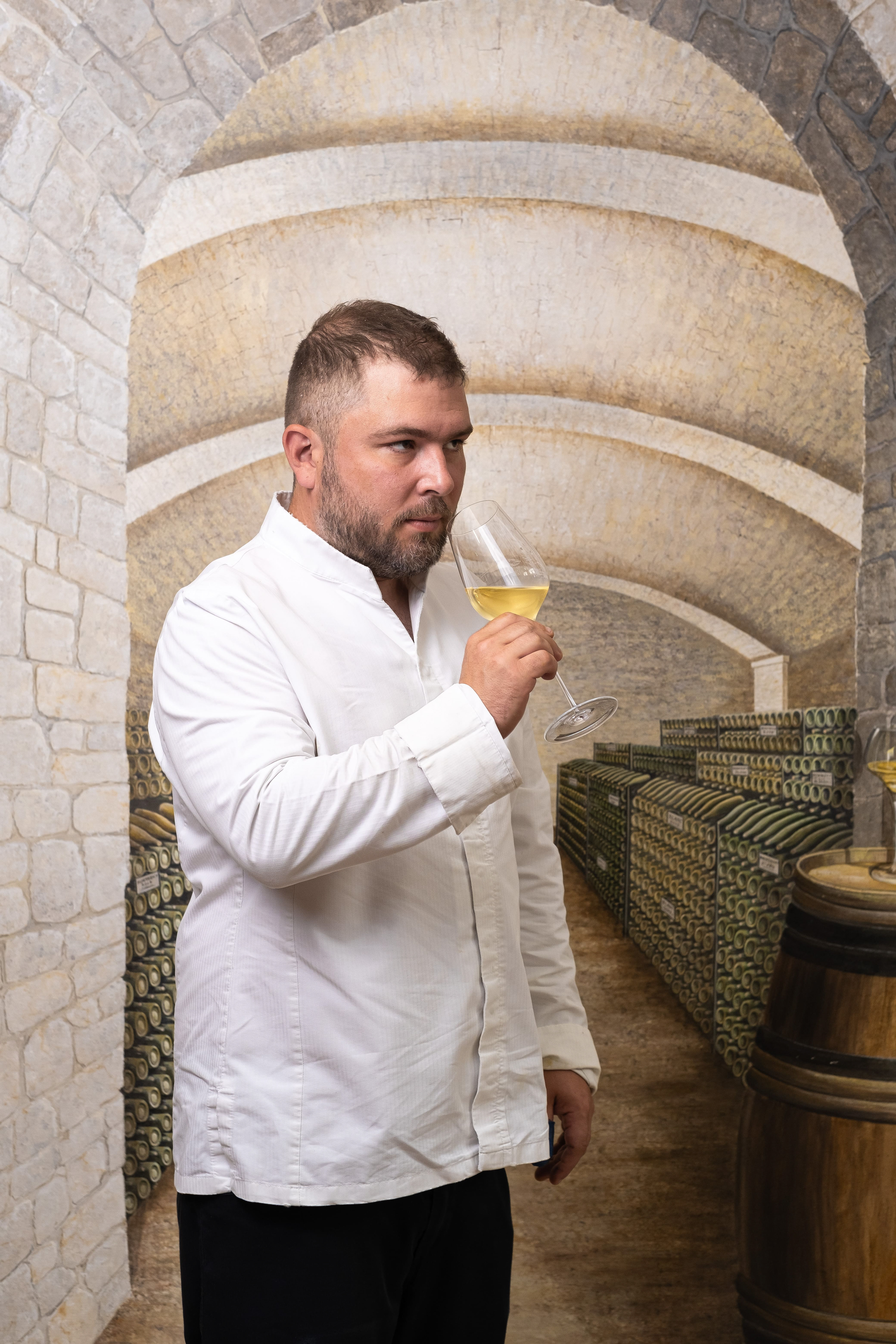 Portrait de Maxime Kowalczyk, chef du restaurant L'Empreinte à Buxy, Saône-et-Loire, Bourgogne, reconnu par Gault et Millau. Il est en train de déguster un verre de vin blanc dans une cave à vin en pierre. Vêtu d'une veste de chef blanche, il incarne l'élégance et le professionnalisme dans un cadre authentique et historique.