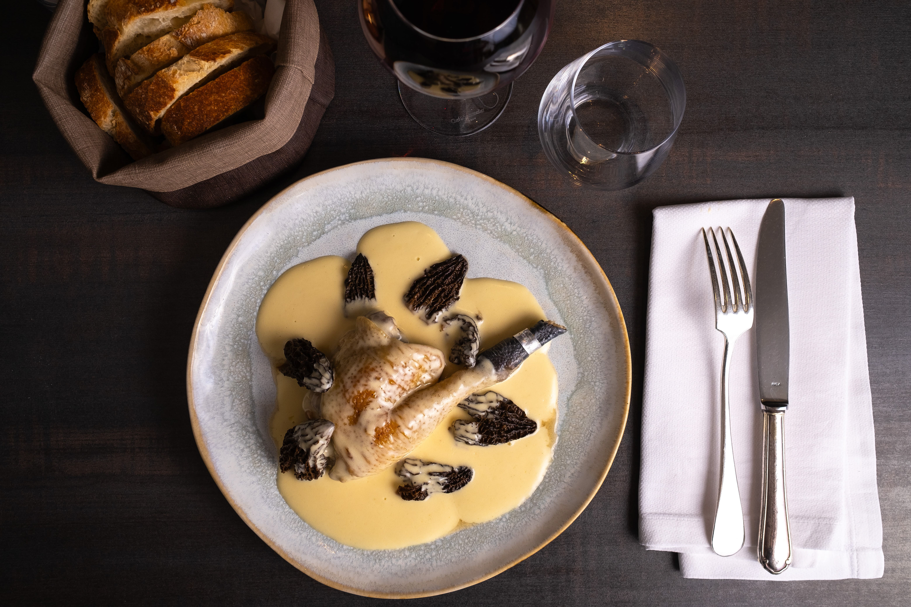 Assiette élégante de volaille de bresse nappée de sauce crémeuse et garnie de morilles, accompagnée d'un panier de tranches de pain frais. La table est dressée avec un verre de vin rouge, un verre d'eau et des couverts en argent posés sur une serviette en tissu blanc. La présentation raffinée du plat et les textures riches des morilles et de la sauce ajoutent une touche gastronomique à cette scène culinaire