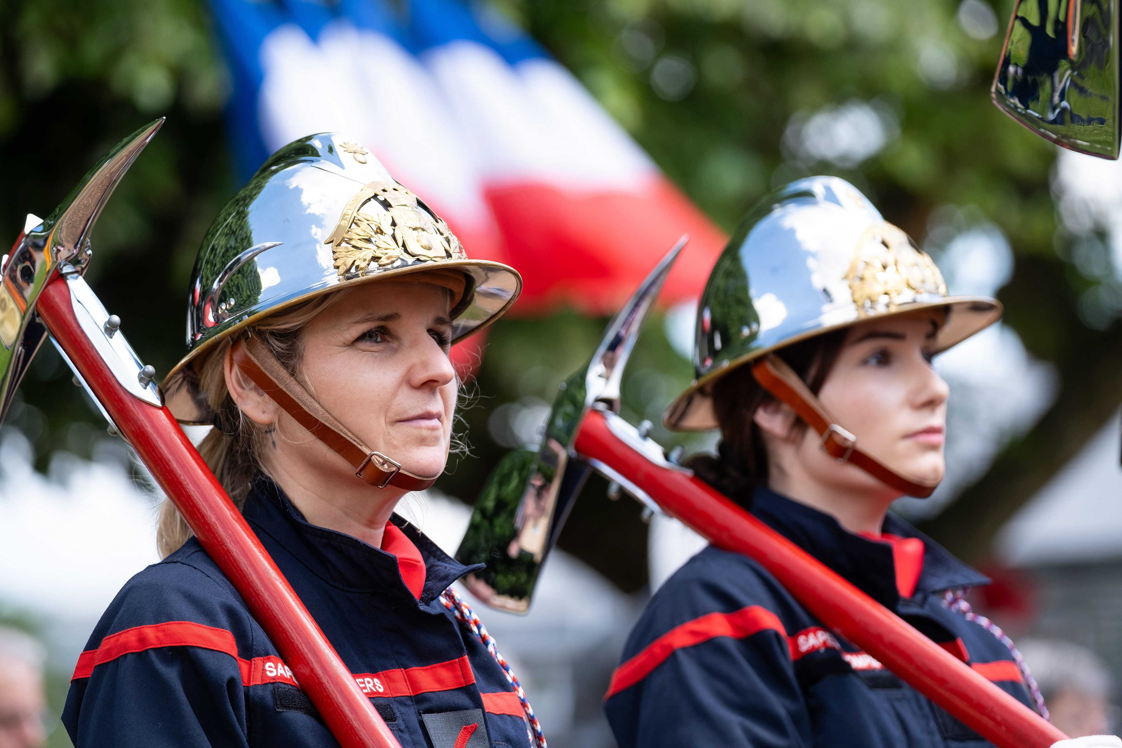 Portrait de deux femmes pompiers en uniforme avec des casques brillants et des haches, lors d'une cérémonie officielle en Saône-et-Loire, Bourgogne, Rhône-Alpes. En arrière-plan, le drapeau français flotte fièrement. Photographie capturée par Etienne Gatheron. Photographie institutionnelle.
