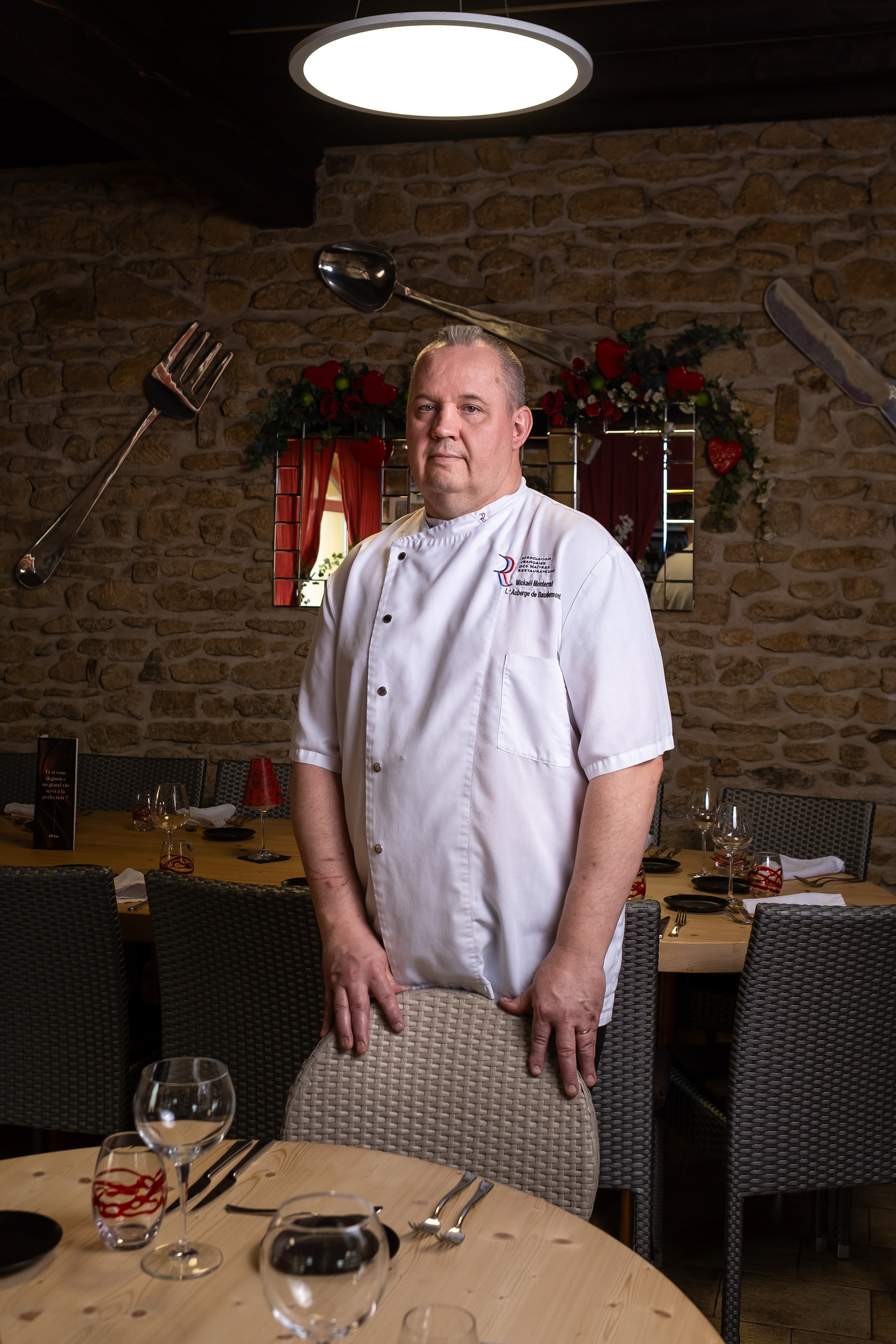 Portrait de Mickaël Monserrat, chef de l'Auberge de Baudemont à Baudemont, Saône-et-Loire, Bourgogne. Debout dans la salle à manger du restaurant, il porte une veste de chef blanche avec le nom de l'établissement brodé. La salle est décorée avec des ustensiles de cuisine géants accrochés aux murs en pierre et des tables élégamment dressées. Cette image met en avant l'atmosphère accueillante et rustique du restaurant, ainsi que le professionnalisme du chef.