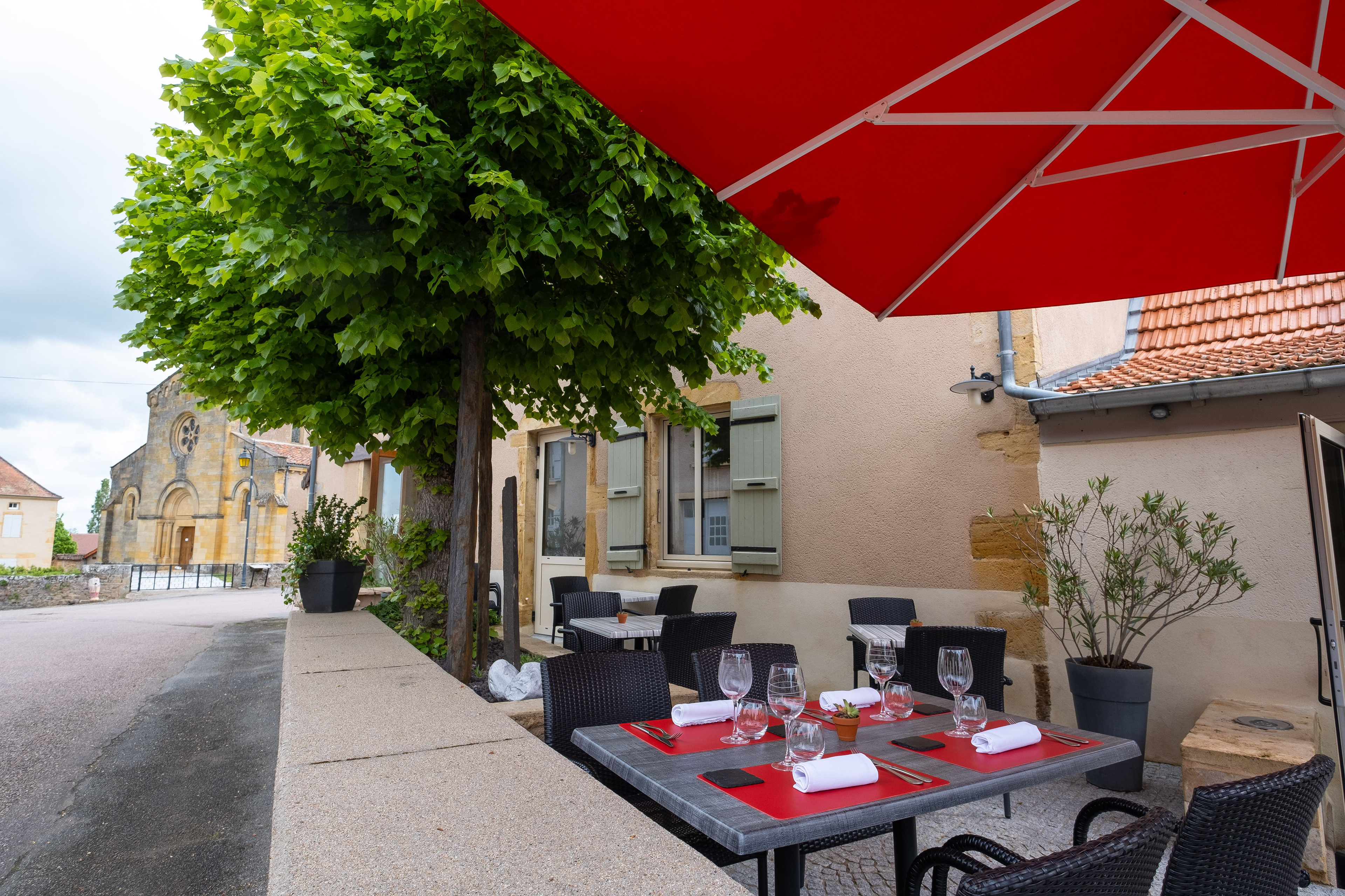 Terrasse de l'Auberge de Briant en Saône-et-Loire, Bourgogne, avec des tables élégamment dressées sous un grand parasol rouge, offrant une vue charmante sur une église ancienne et des bâtiments traditionnels, dans un cadre pittoresque et accueillant.