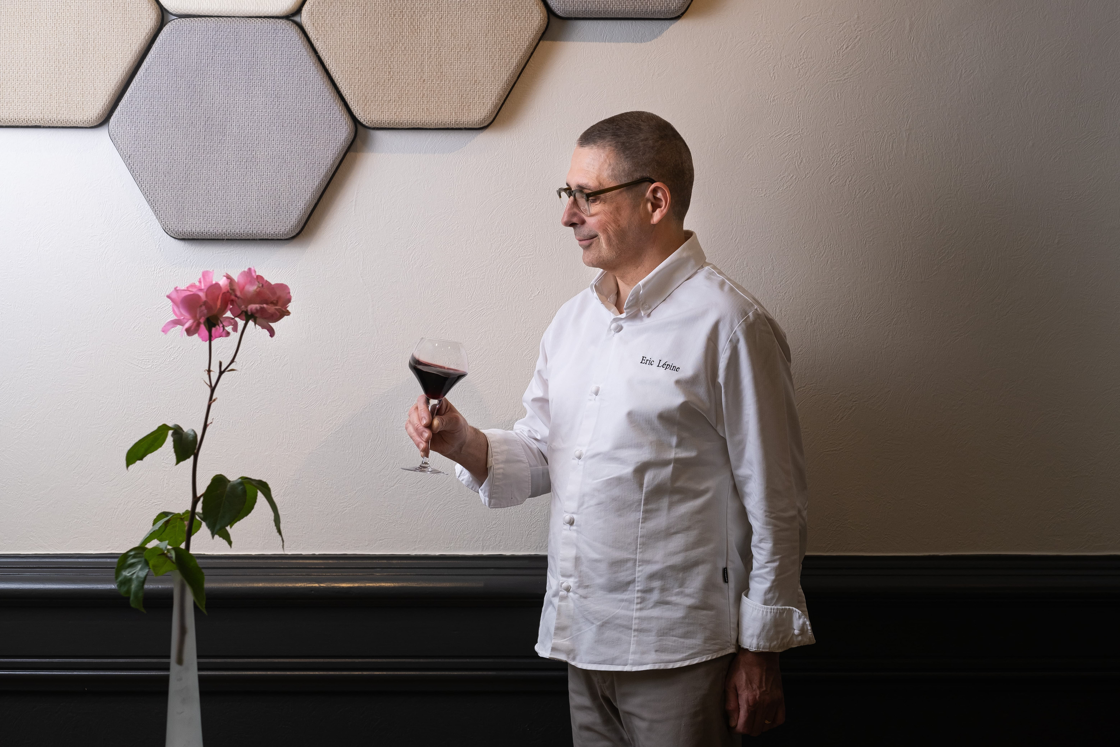 Portrait d'Éric Lépine, chef du Restaurant Emile Job à Saint-Sernin, Saône-et-Loire, Bourgogne, tenant un verre de vin rouge. Il est vêtu d'une veste de chef blanche personnalisée avec son nom. À l'arrière-plan, une décoration murale hexagonale et un vase avec des fleurs roses ajoutent une touche esthétique. Cette image capture l'élégance et la sophistication du chef dans un environnement raffiné.