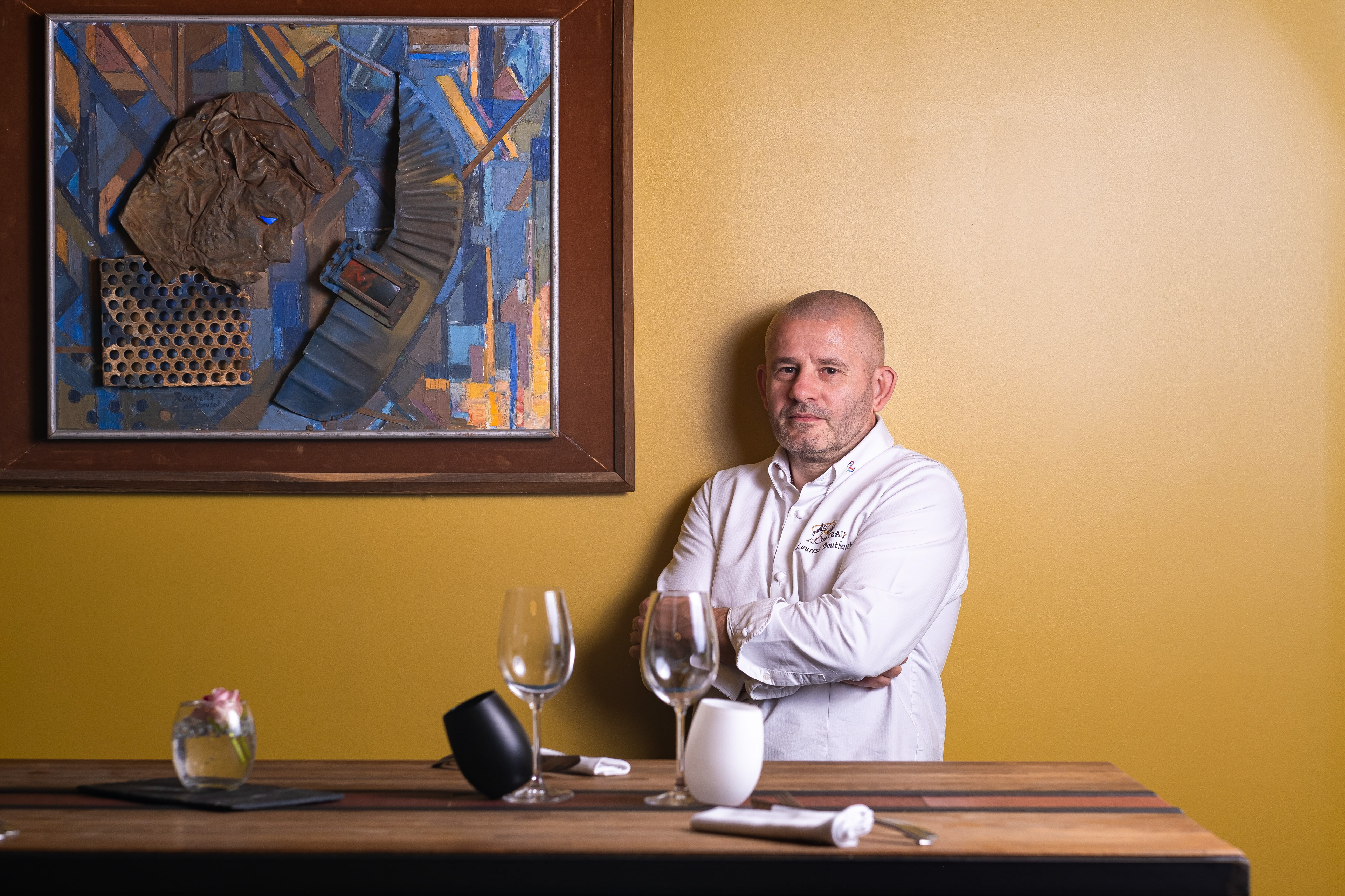 Portrait de Laurent Bouthenet, chef du Restaurant du Château à Saint-Sernin, Saône-et-Loire, Bourgogne, debout devant une table avec les bras croisés, vêtu d'une veste de chef blanche personnalisée. En arrière-plan, une peinture murale abstraite aux couleurs vives et un mur jaune créent un contraste saisissant. Sur la table, des verres à vin, une coupe noire et blanche, et une petite composition florale renforcent l'élégance de l'établissement.