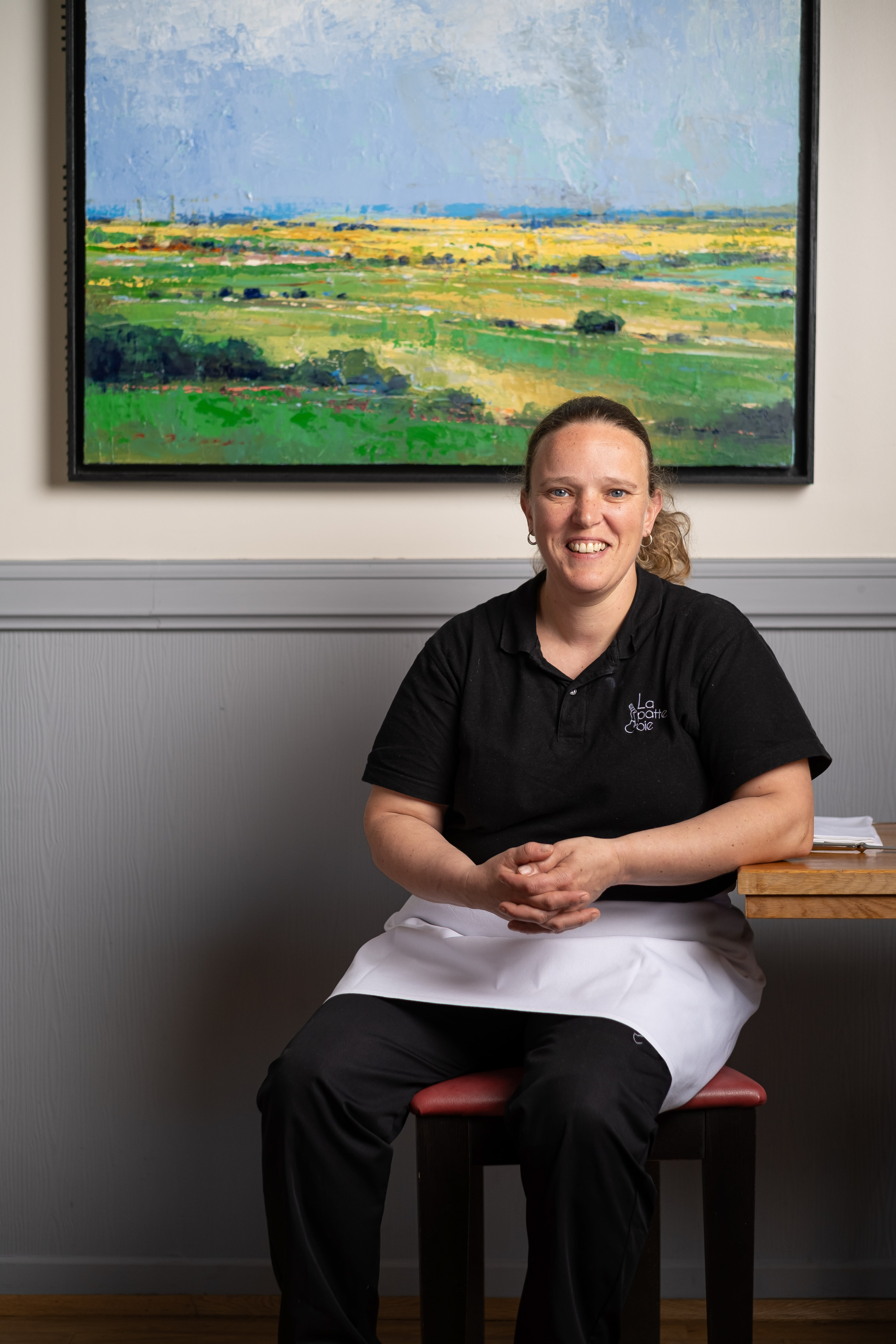 Portrait d'Estelle, chef de cuisine au Restaurant La Patte d'Oie à Davayé, Saône-et-Loire, Bourgogne, assise devant une peinture de paysage colorée. Elle porte un polo noir et un tablier blanc, et affiche un sourire chaleureux. Cette image capture l'ambiance accueillante et artistique du restaurant, mettant en avant la personnalité et le professionnalisme du chef.