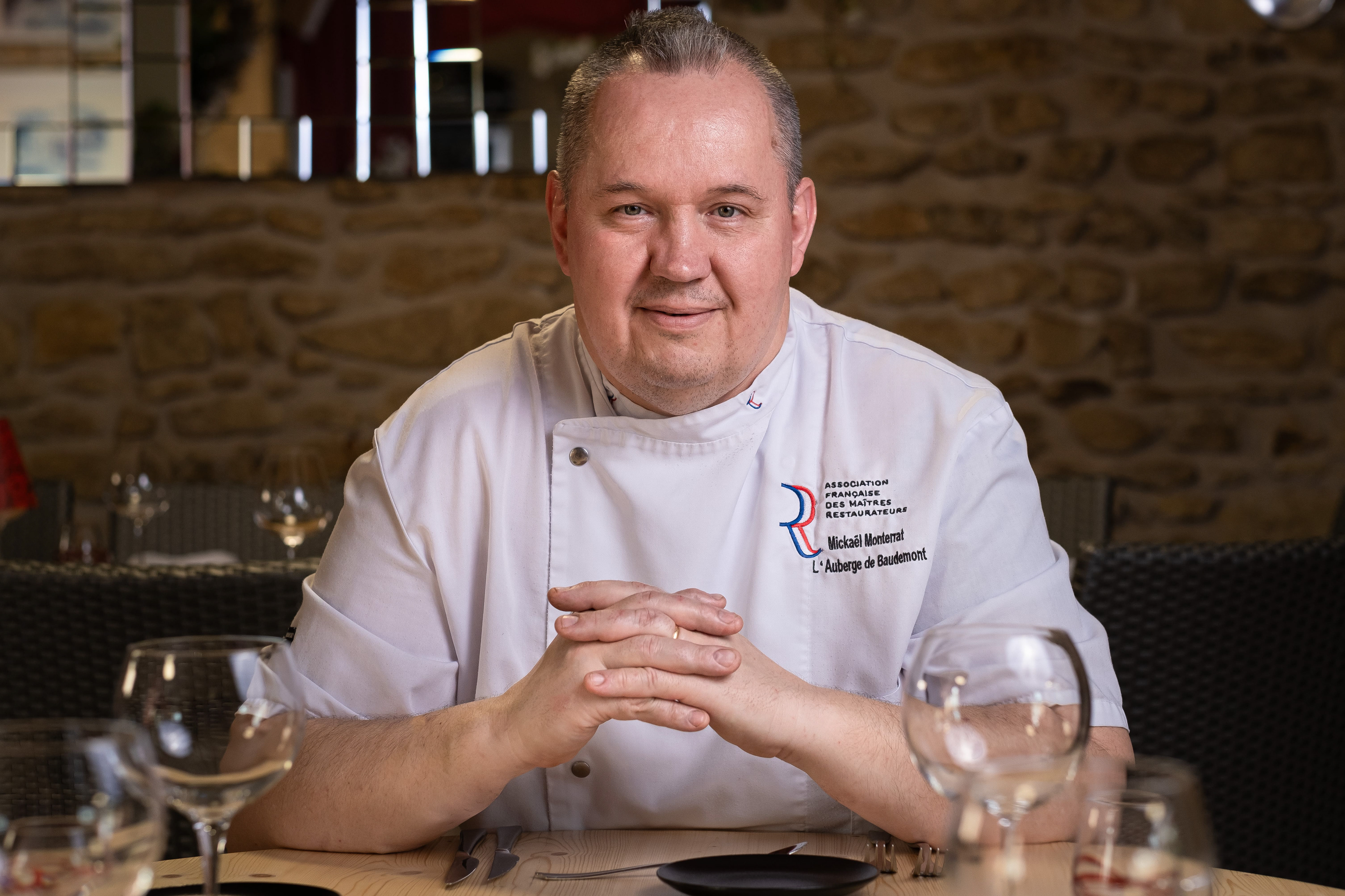Portrait de Mickaël Monterratt, chef de l'Auberge de Baudemont à Baudemont, Saône-et-Loire, Bourgogne, reconnu par l'Association Française des Maîtres Restaurateurs. Assis à une table élégamment dressée dans la salle à manger du restaurant, il porte une veste de chef blanche avec son nom et le logo de l'association. La salle est décorée avec des murs en pierre, renforçant l'ambiance rustique et chaleureuse du restaurant.
