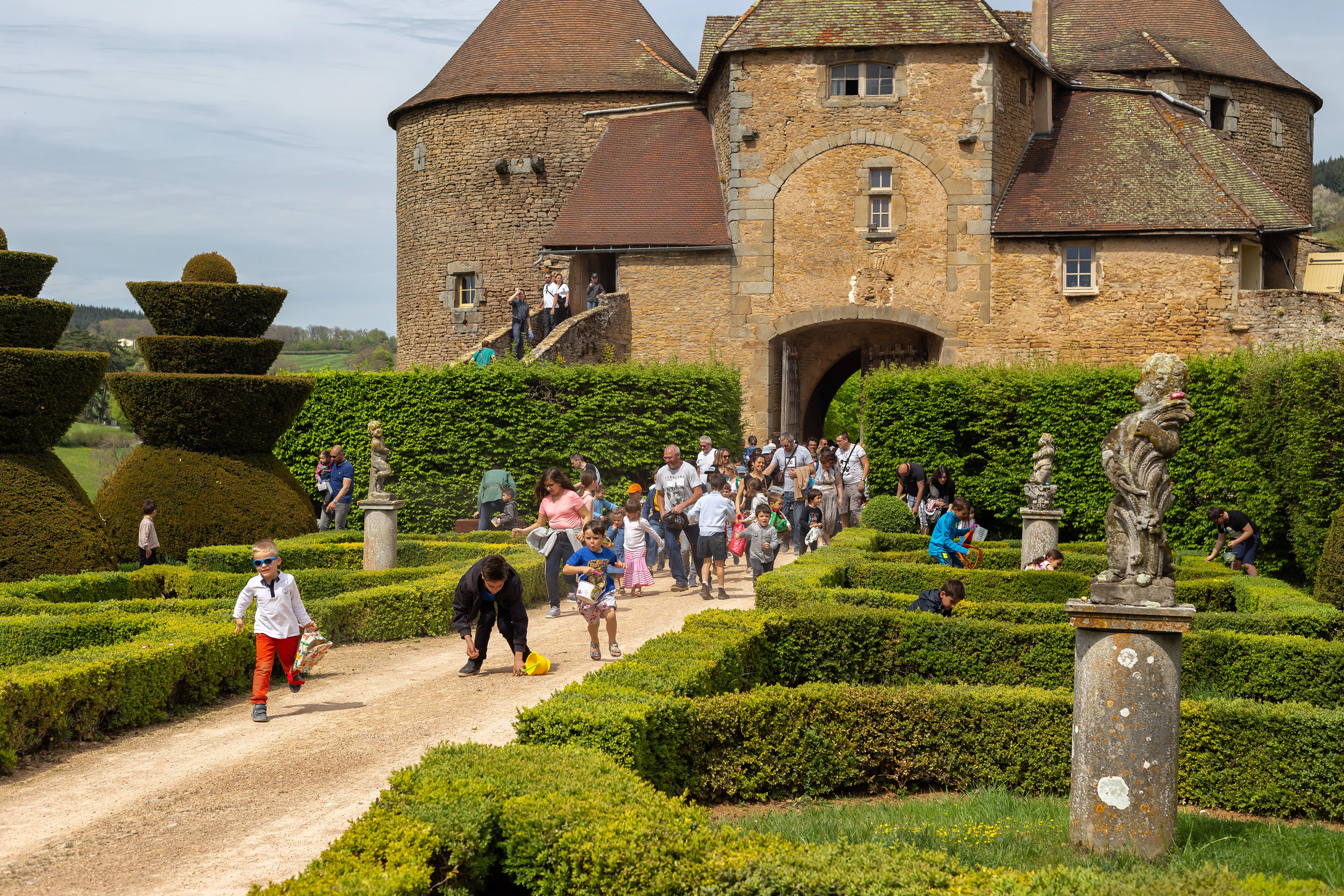 Visiteurs explorant les jardins et structures historiques de la Citadelle de Berzé le Châtel en Saône-et-Loire, Bourgogne. Scène vivante avec enfants et familles sous ciel partiellement nuageux. Patrimoine culturel et attrait touristique. Photographie touristique par Etienne Gatheron.