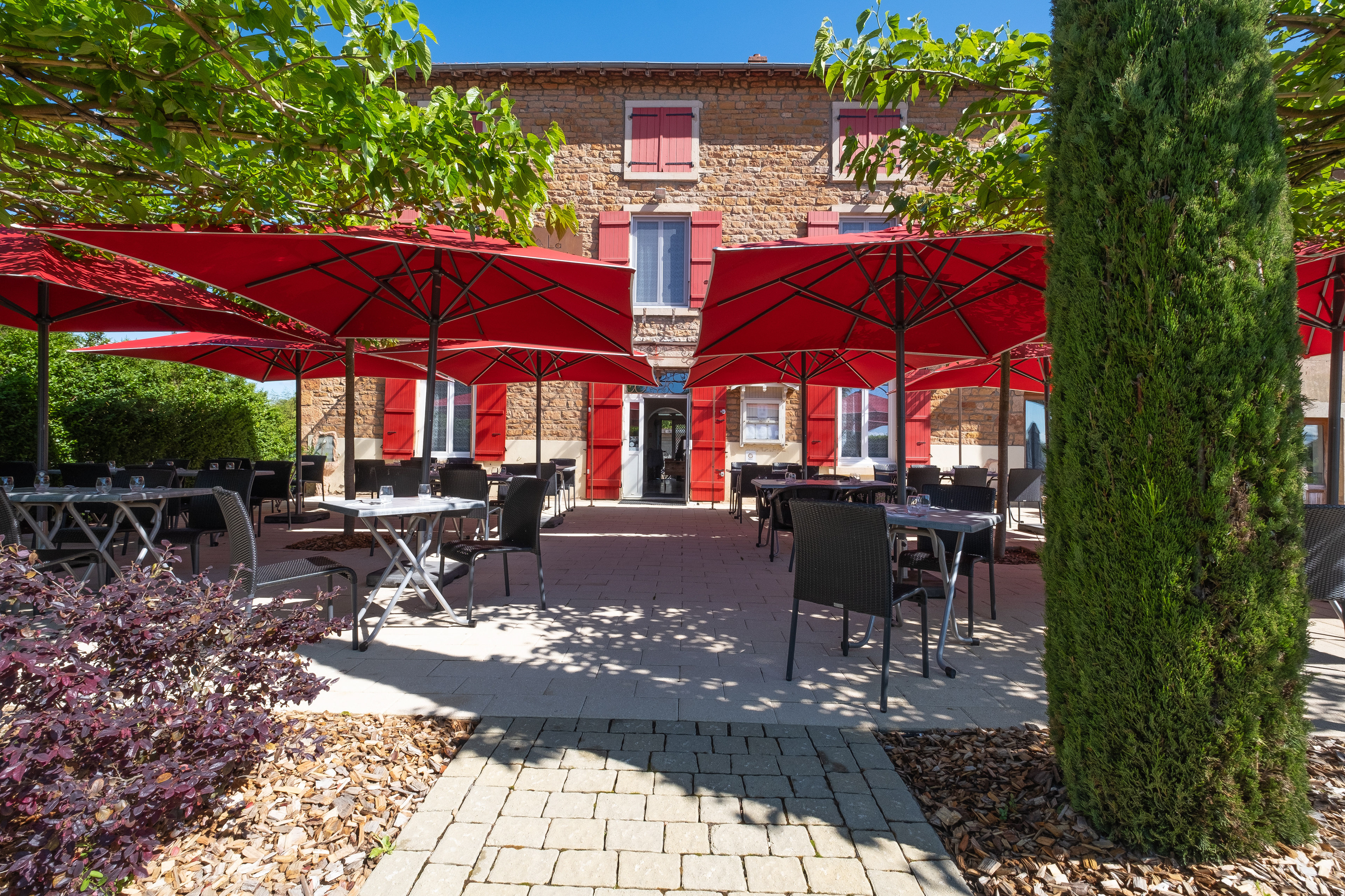 Terrasse de La Patte d'Oie à Davayé en Saône-et-Loire, Bourgogne, montrant des tables élégamment dressées sous de grands parasols rouges, avec une façade en pierre et des volets rouges en arrière-plan, offrant un cadre charmant et accueillant pour un repas en plein air.