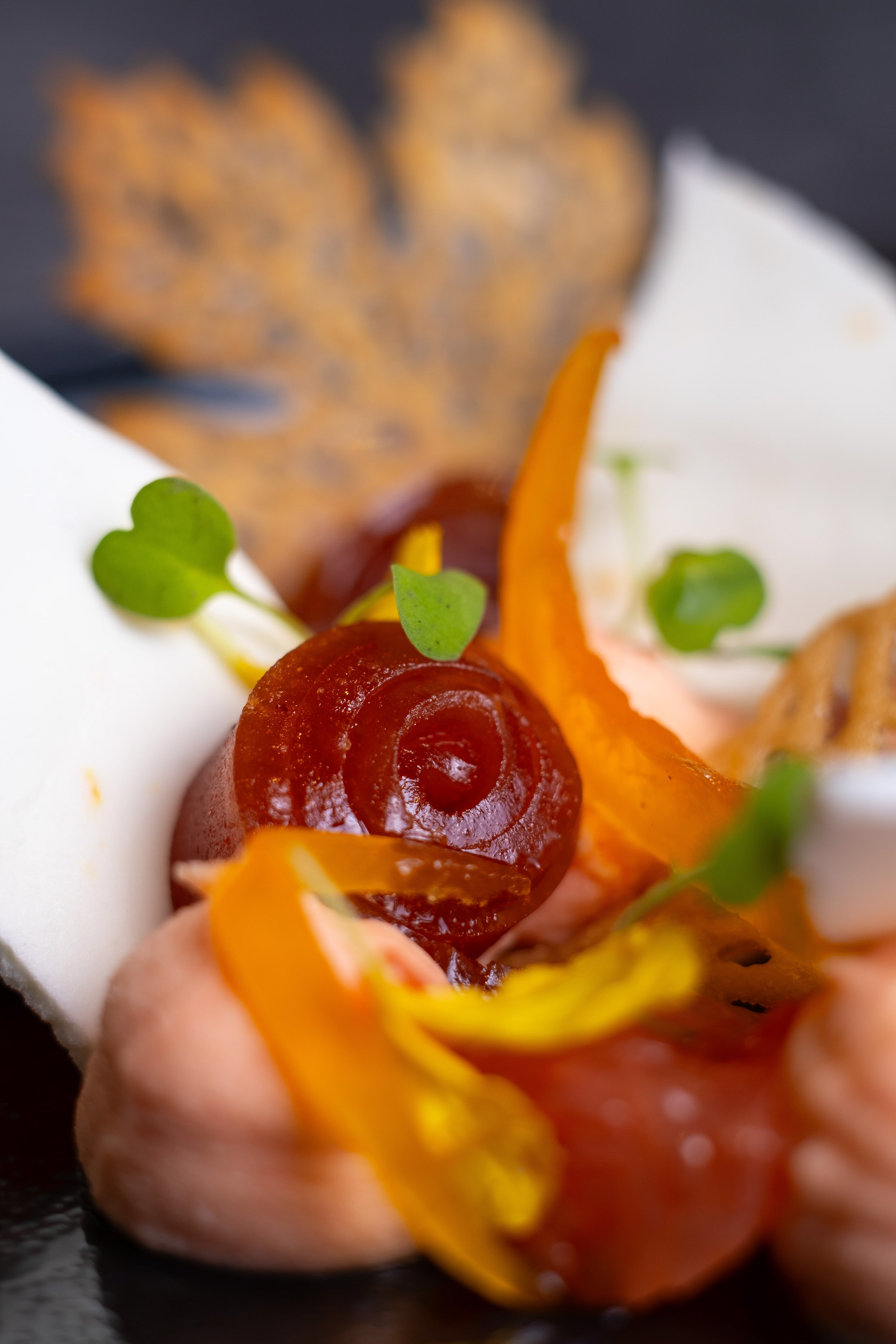 Gros plan sur une préparation de dessert gastronomique sophistiquée avec des éléments artistiques tels que des spirales de gelée rouge, des pétales d'orange translucide, des herbes fraîches et des feuilles croquantes. La présentation soignée met en valeur la texture et les couleurs vibrantes des ingrédients, créant une composition visuelle attrayante qui souligne l'attention aux détails et la créativité culinaire du chef.