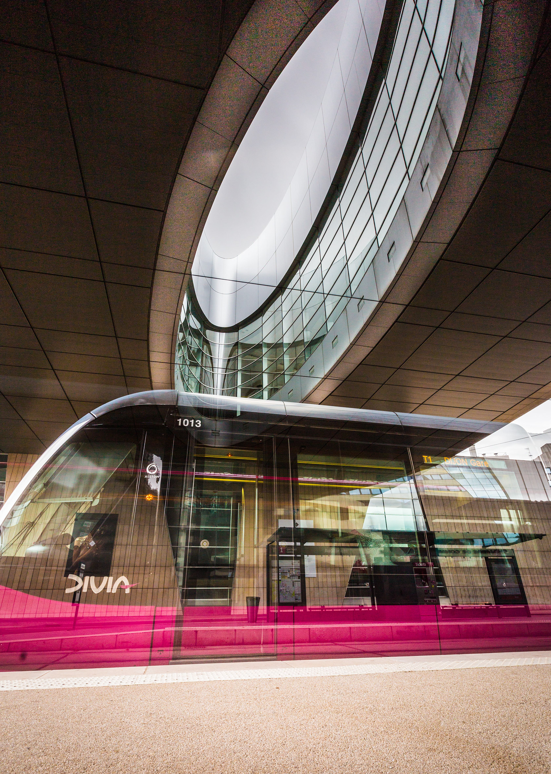 Tramway moderne de la ligne Divia à Dijon : Tramway stationné sous une structure architecturale incurvée avec des façades en verre et un ciel gris visible à travers les ouvertures. Photographie par Etienne Gatheron, expert en architecture moderne.