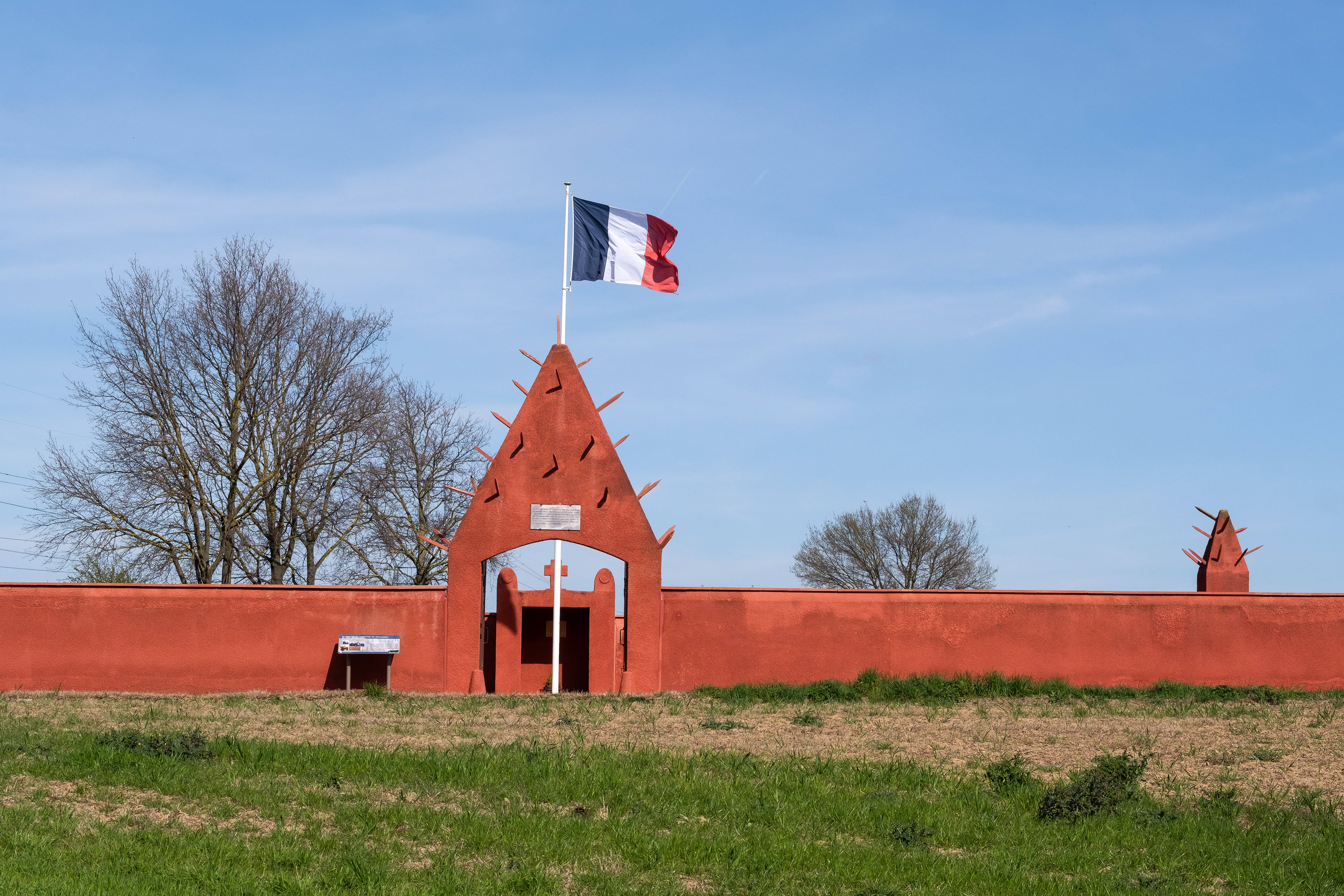 Mur rouge distinctif, stèles blanches et plaque commémorative au Cimetière Militaire Tata de Chasselay. Hommage aux tirailleurs sénégalais de la Seconde Guerre mondiale. Patrimoine mémoriel et attrait touristique. Photo par Etienne Gatheron, spécialiste des sites historiques, patrimoine culturel et tourisme mémoriel.
