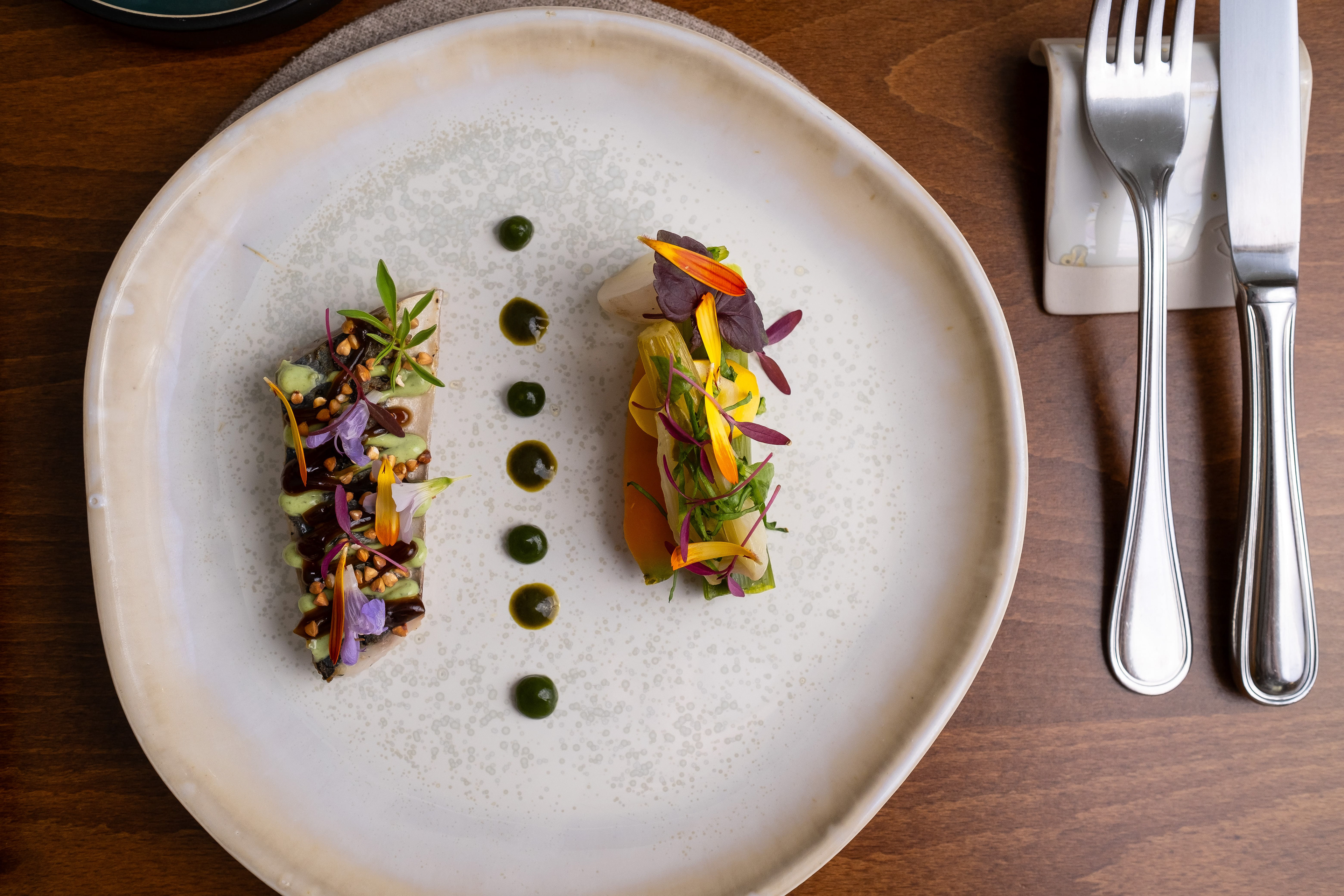 Vue de dessus d'une assiette gastronomique composée de deux préparations artistiques de poisson ornées de fleurs comestibles et de jeunes pousses, disposées en symétrie et accompagnées de gouttes de sauce verte. Les détails colorés des fleurs et la présentation soignée sur une assiette en céramique ajoutent une touche d'élégance à cette création culinaire. À côté de l'assiette, des couverts en acier inoxydable sont placés sur un repose-couverts en céramique blanche