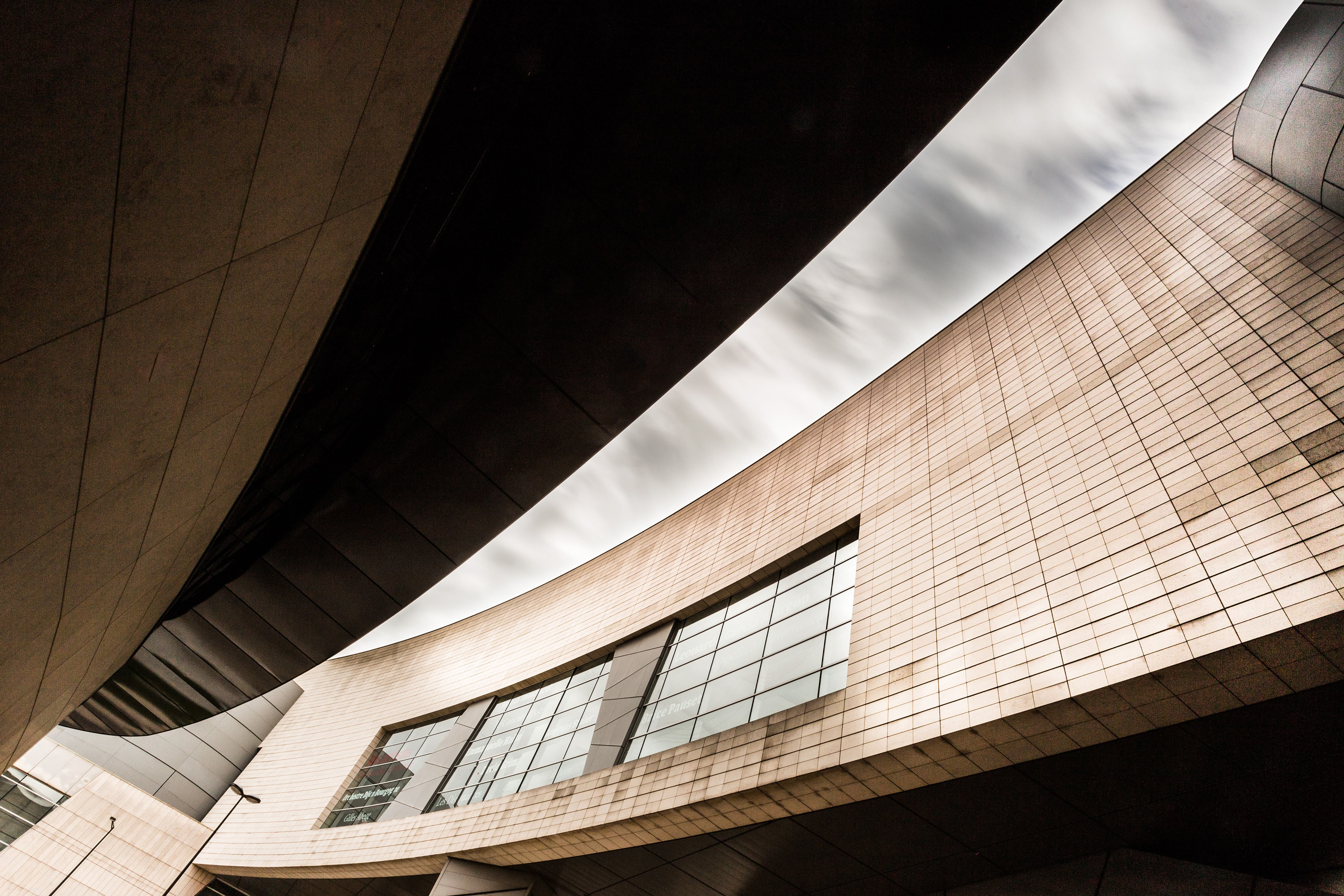 Vue panoramique de l'auditorium de Dijon : Angle unique montrant la courbe fluide du bâtiment et les jeux de lumière et d'ombre, avec un ciel nuageux en arrière-plan. Photographie par Etienne Gatheron, spécialiste en architecture.