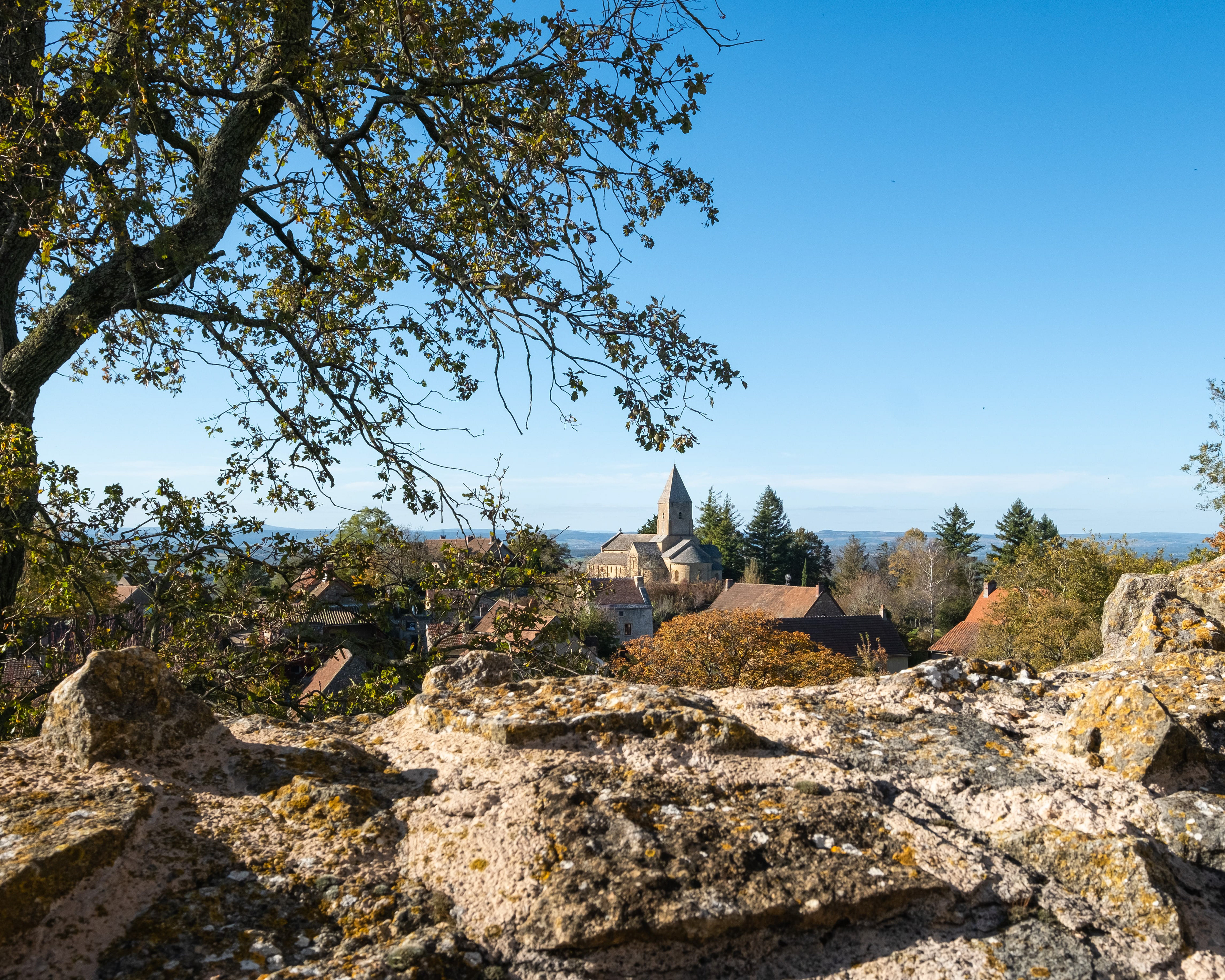 Château de Brancion entouré de branches d'arbres et formations rocheuses, avec clocher de l'église romane, maisons traditionnelles et feuillage automnal. Patrimoine architectural et beauté naturelle de Saône-et-Loire, Bourgogne. Photographie touristique par Etienne Gatheron.