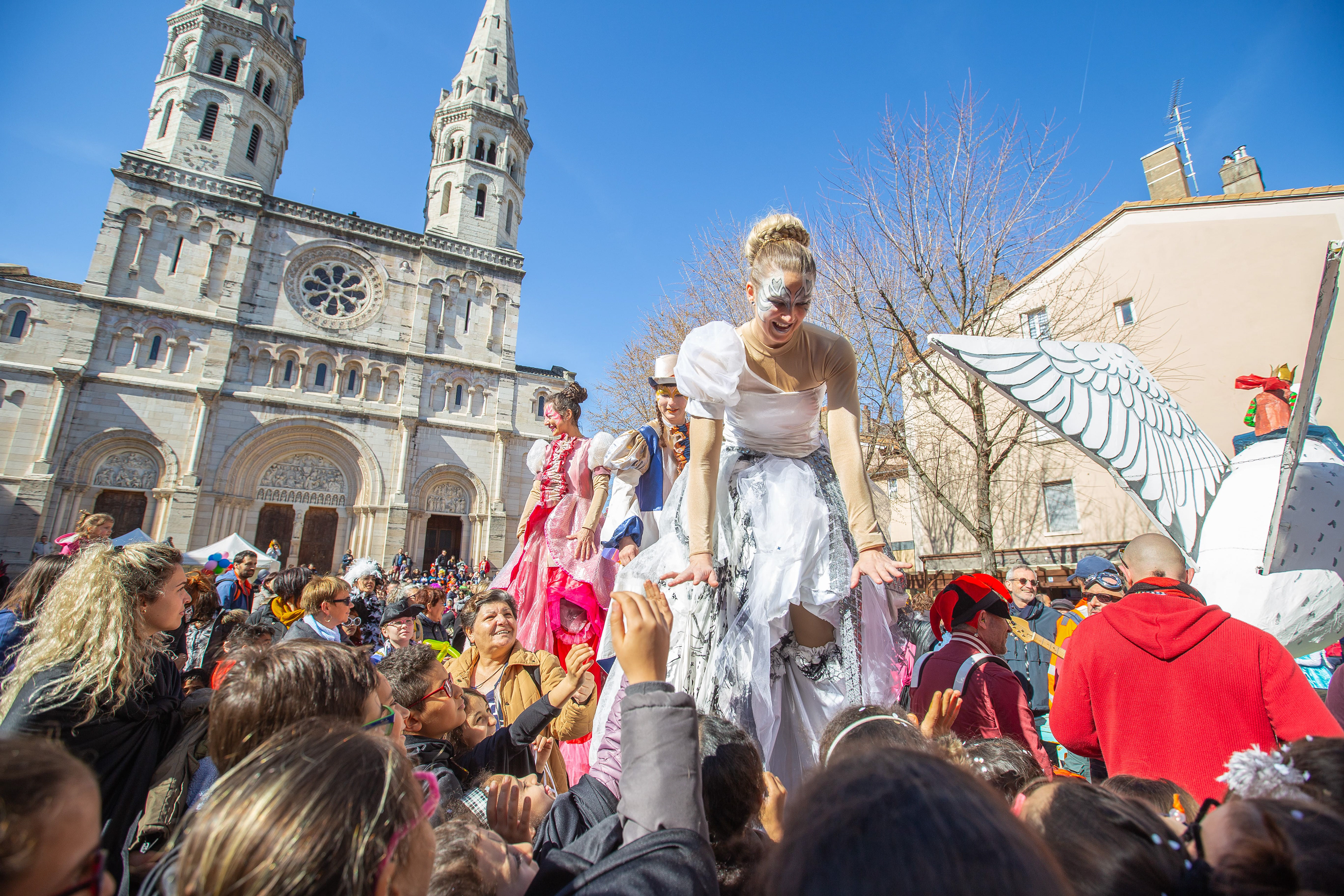 Performers costumés sur échasses interagissant avec la foule lors d'un carnaval festif devant la cathédrale de Mâcon, Saône-et-Loire, Bourgogne, Rhône-Alpes, sous un ciel bleu clair. Photographie capturée par Etienne Gatheron. Photographie institutionnelle.