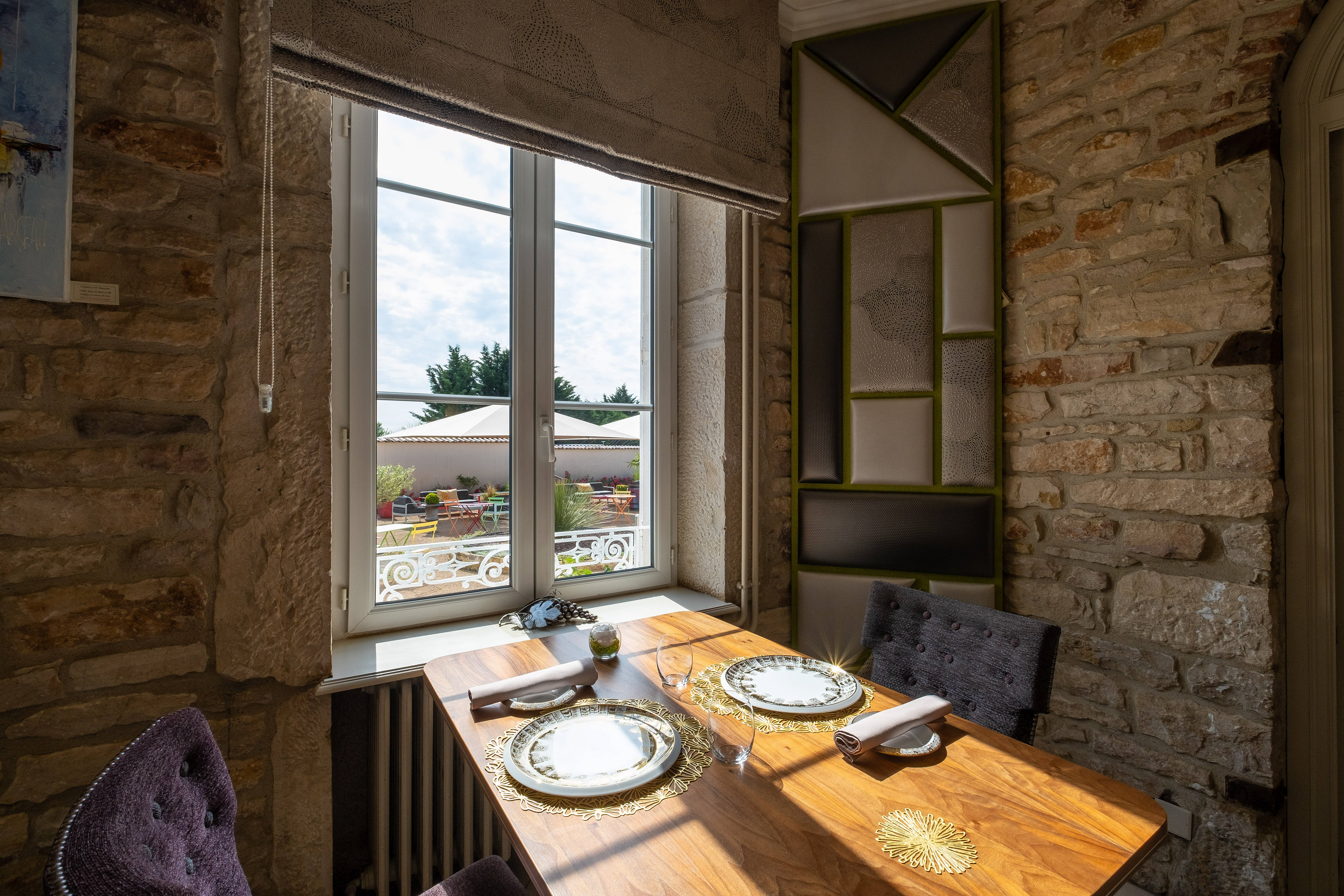 Table élégamment dressée à La Marande à Montbellet en Saône-et-Loire, Bourgogne, avec des assiettes dorées et des serviettes blanches, située près d'une grande fenêtre offrant une vue sur une terrasse colorée et des jardins, dans un cadre intérieur en pierre.