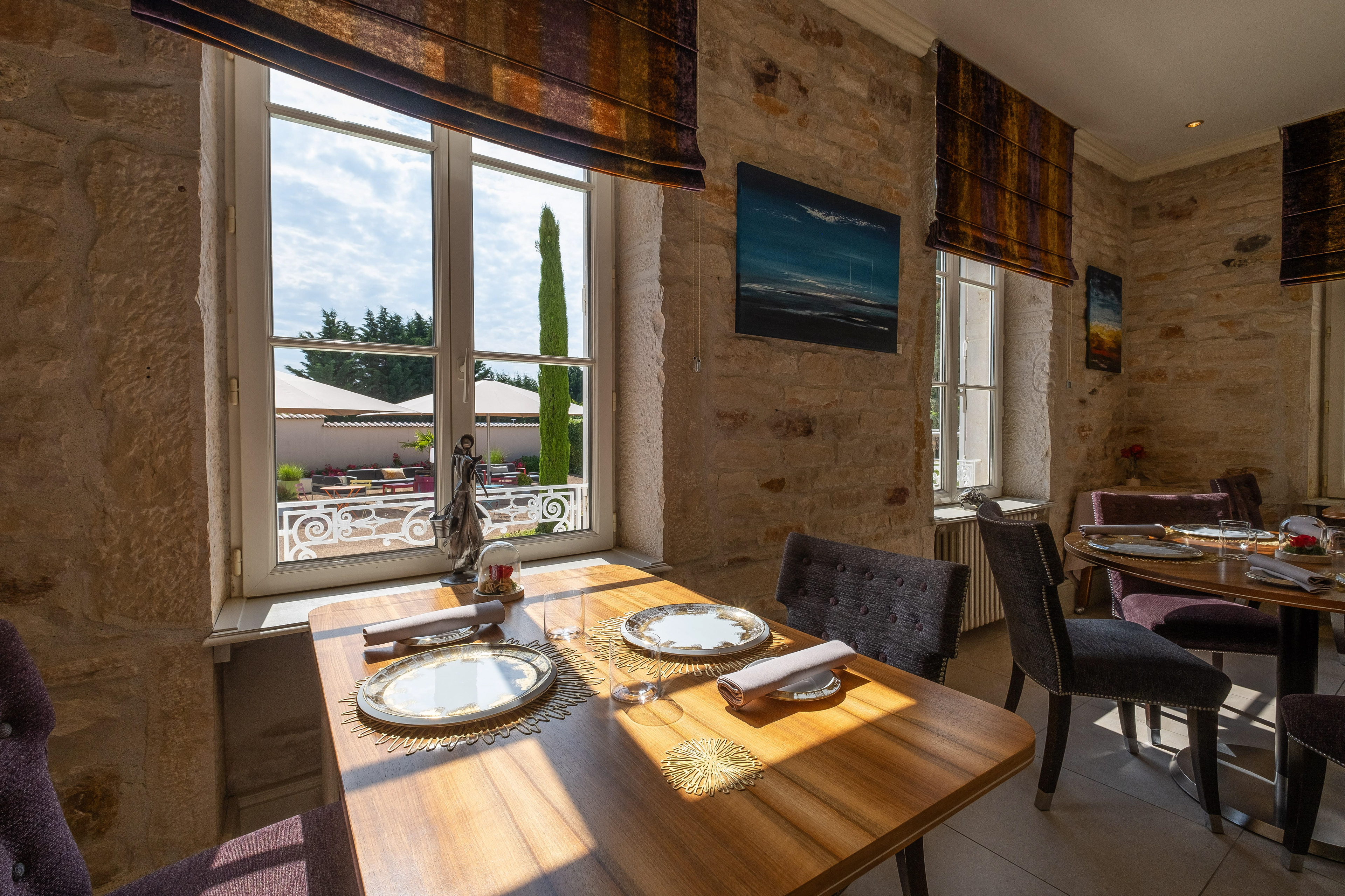 Salle à manger de La Marande à Montbellet en Saône-et-Loire, Bourgogne, montrant une table élégamment dressée avec des assiettes dorées et des serviettes blanches, dans un cadre lumineux avec des murs en pierre et une grande fenêtre offrant une vue sur une terrasse ensoleillée et des jardins.