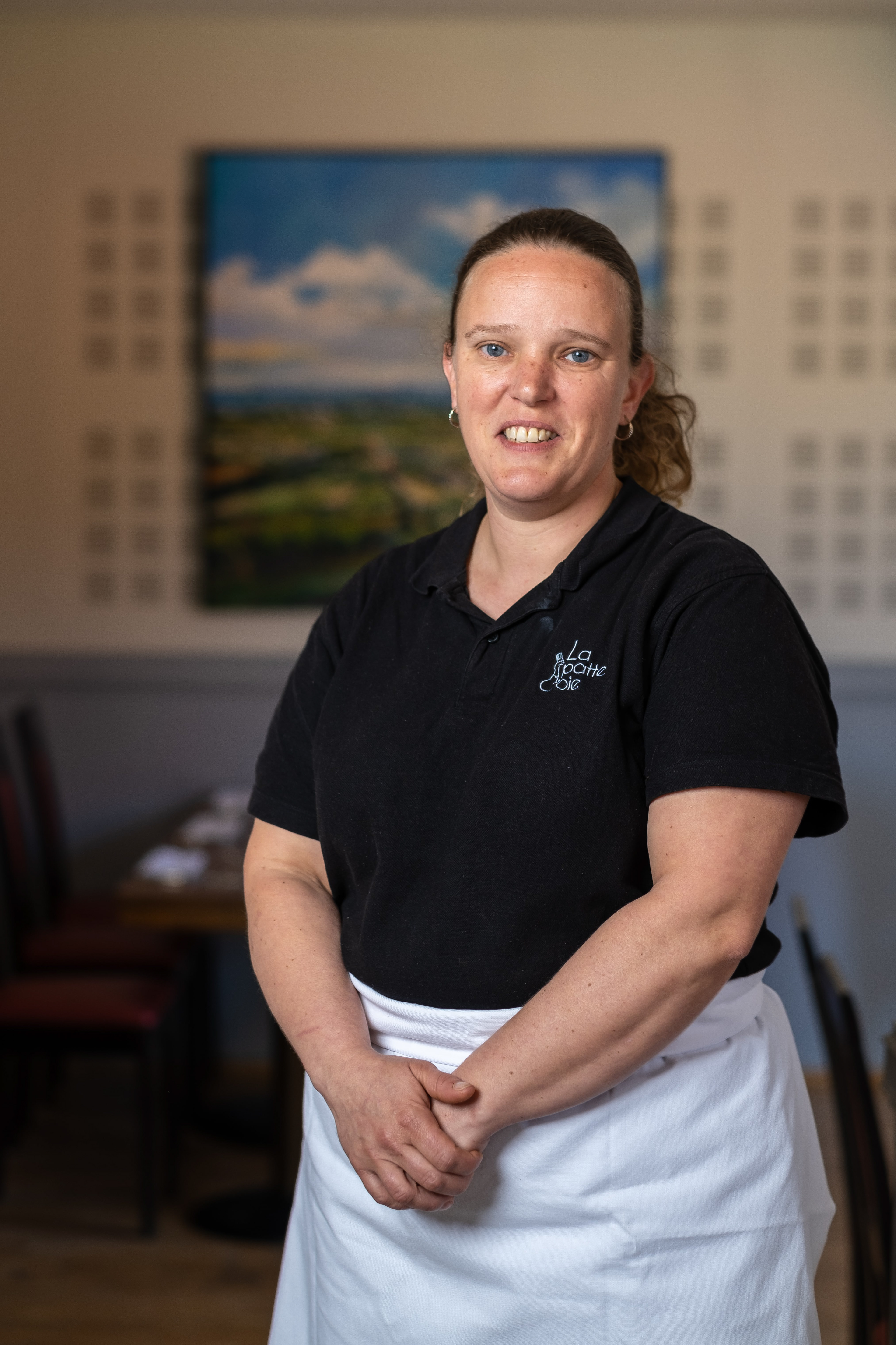 Portrait d'Estelle, chef de cuisine au Restaurant La Patte d'Oie à Davayé, Saône-et-Loire, Bourgogne, habillée d'un polo noir et d'un tablier blanc. Elle se tient debout dans la salle à manger du restaurant avec un sourire accueillant. En arrière-plan, une peinture de paysage ajoute une touche artistique à l'ambiance chaleureuse de l'établissement.