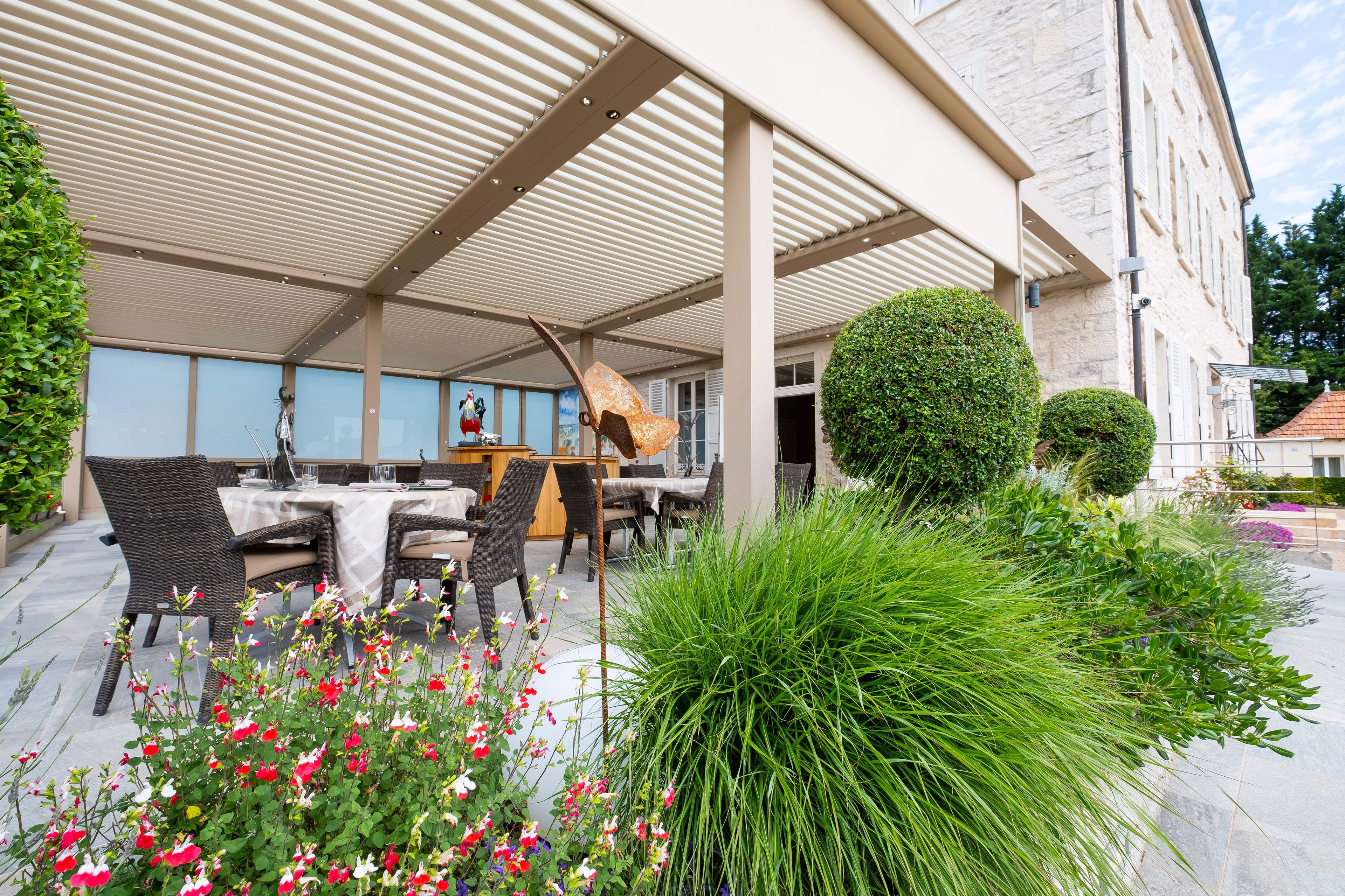 Terrasse de La Marande à Montbellet en Saône-et-Loire, Bourgogne, avec des tables élégamment dressées sous une pergola moderne, entourée de plantes fleuries et d'arbustes taillés, offrant un cadre agréable et verdoyant pour un repas en plein air.