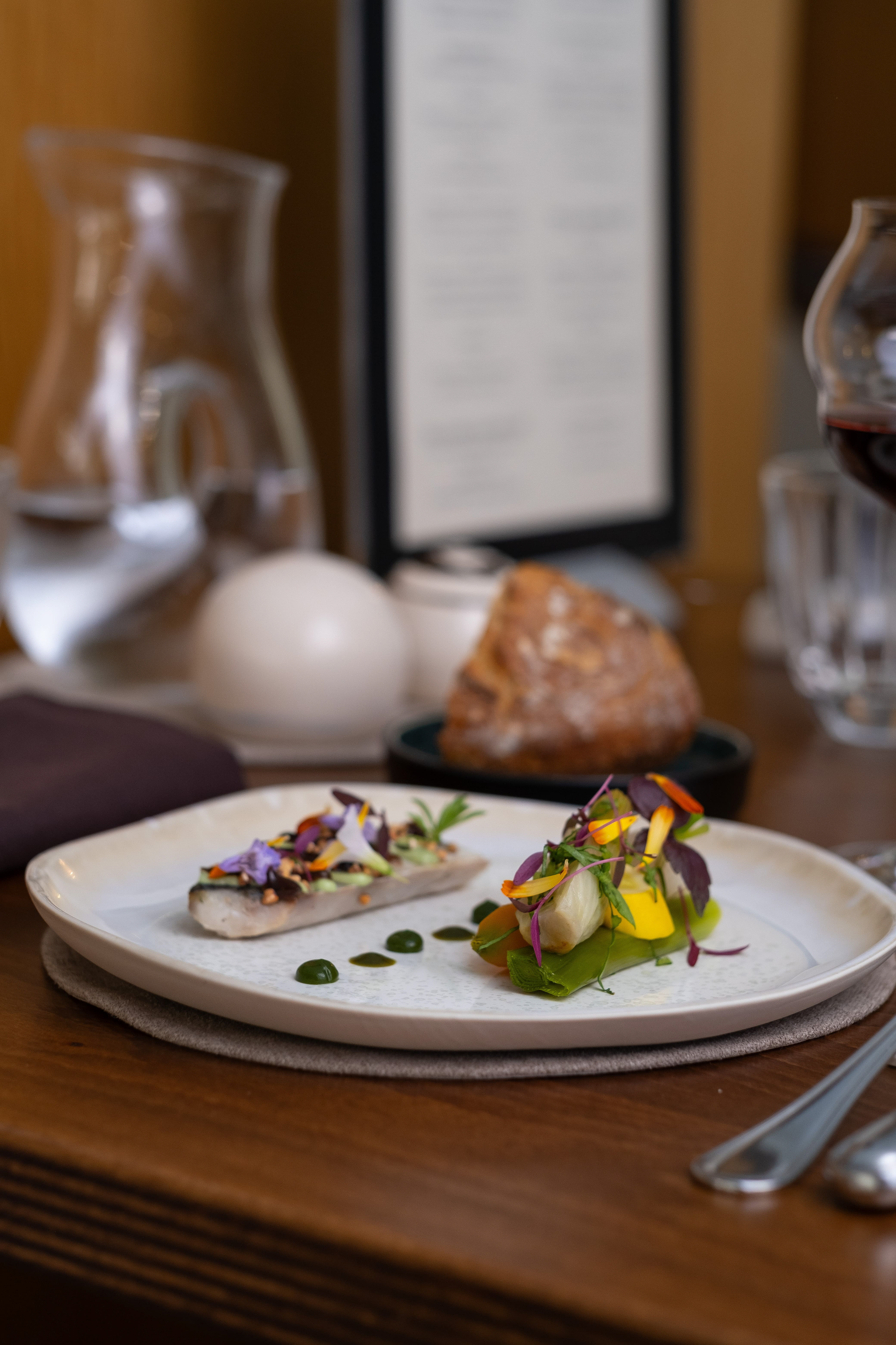 Assiette gastronomique composée d'une préparation de poisson ornée de fleurs comestibles et d'un accompagnement de légumes colorés. La table est décorée avec un verre de vin rouge, une carafe d'eau, un panier de pain artisanal et un menu en arrière-plan. Les couverts en acier inoxydable ajoutent une touche d'élégance à cette présentation culinaire raffinée