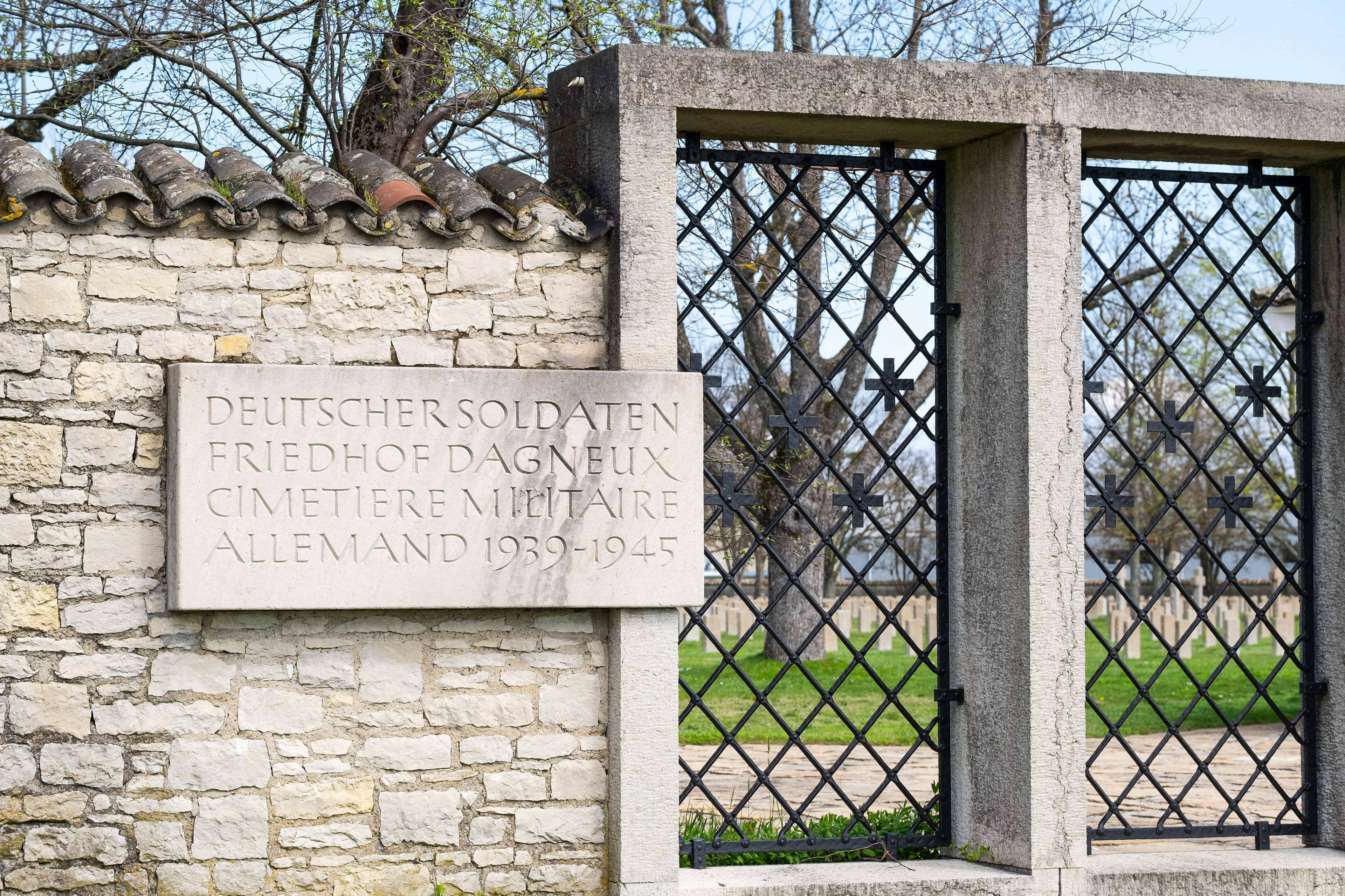 Entrée du Cimetière Militaire Allemand de Dagneux près de Lyon, avec mur en pierre, plaque commémorative et grilles en fer forgé. Rangées de tombes visibles à l'intérieur, illustrant le patrimoine mémoriel et touristique du site. Photo par Etienne Gatheron, spécialiste des sites historiques, patrimoine culturel et tourisme mémoriel.