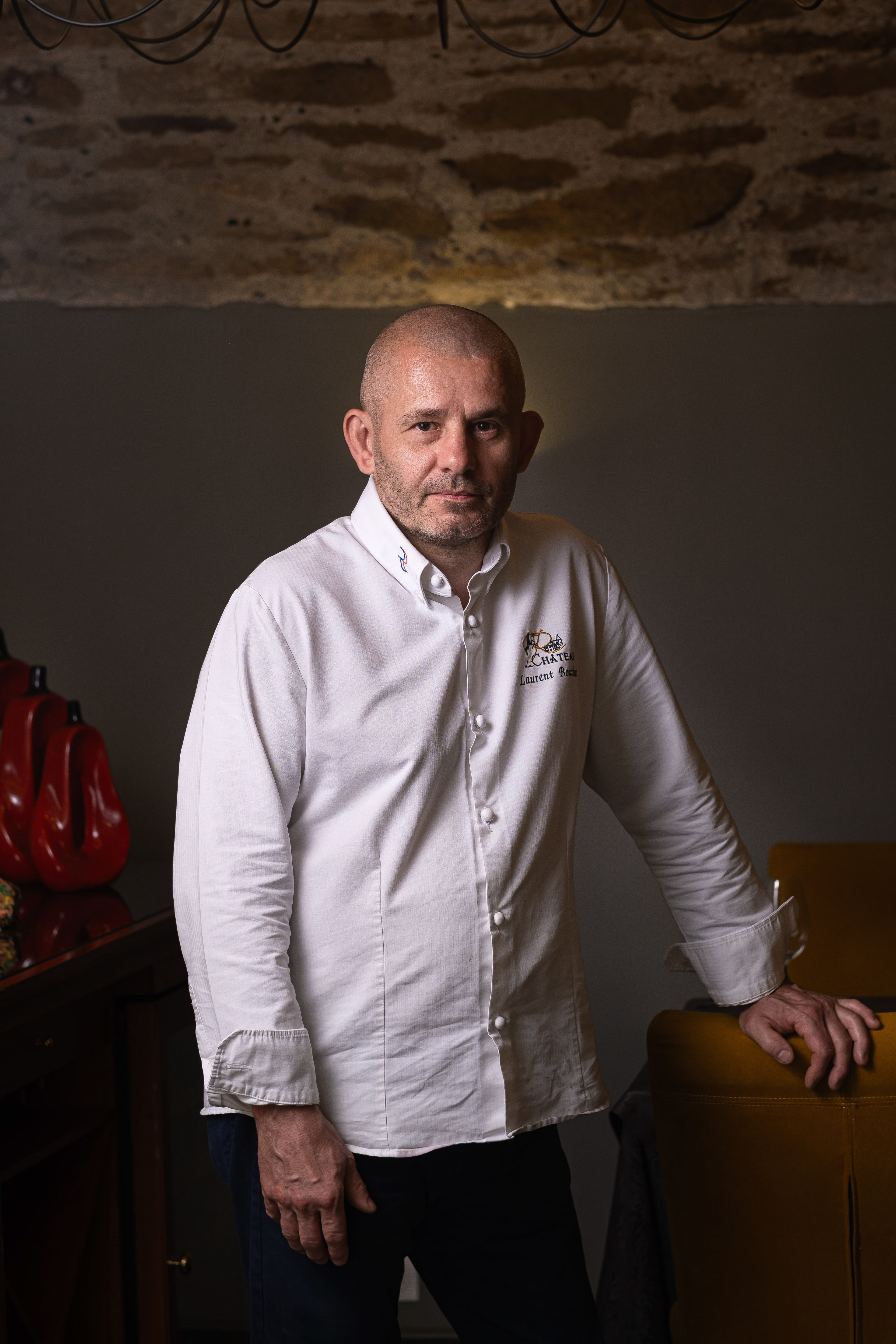 Portrait de Laurent Bouthenet, chef du Restaurant du Château à Saint-Sernin, Saône-et-Loire, Bourgogne, debout devant un mur de pierre, vêtu d'une veste de chef blanche avec son nom et le logo du restaurant. Il est appuyé sur une chaise en cuir jaune, ajoutant une touche de sophistication à la scène. En arrière-plan, des éléments décoratifs rouges apportent une touche de couleur vive.