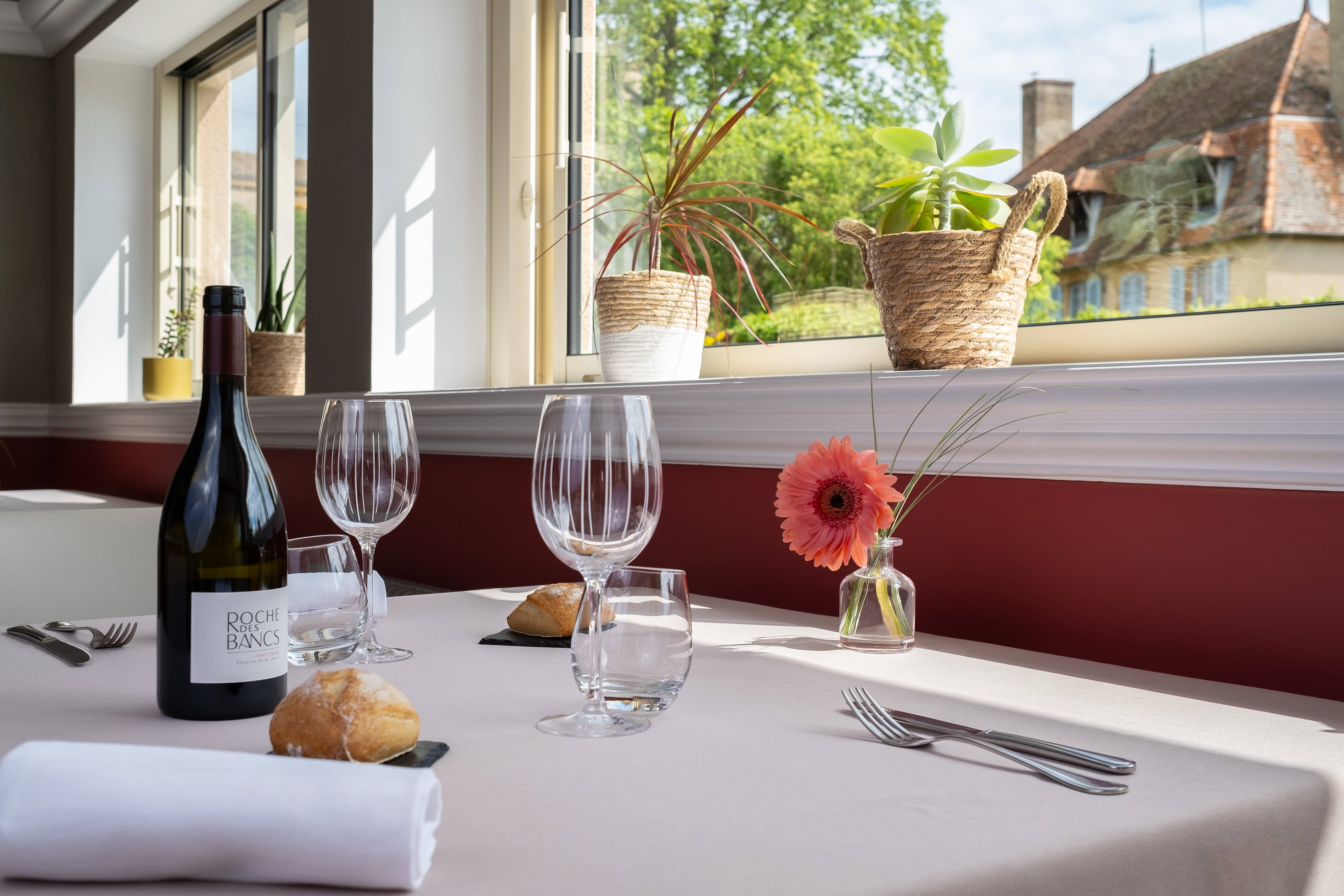 Photo culinaire de l'Auberge de Briant en Saône-et-Loire, Bourgogne, montrant une bouteille de vin Roche des Bancs, des verres à vin, et une fleur dans un cadre lumineux avec vue sur des plantes et des bâtiments traditionnels.