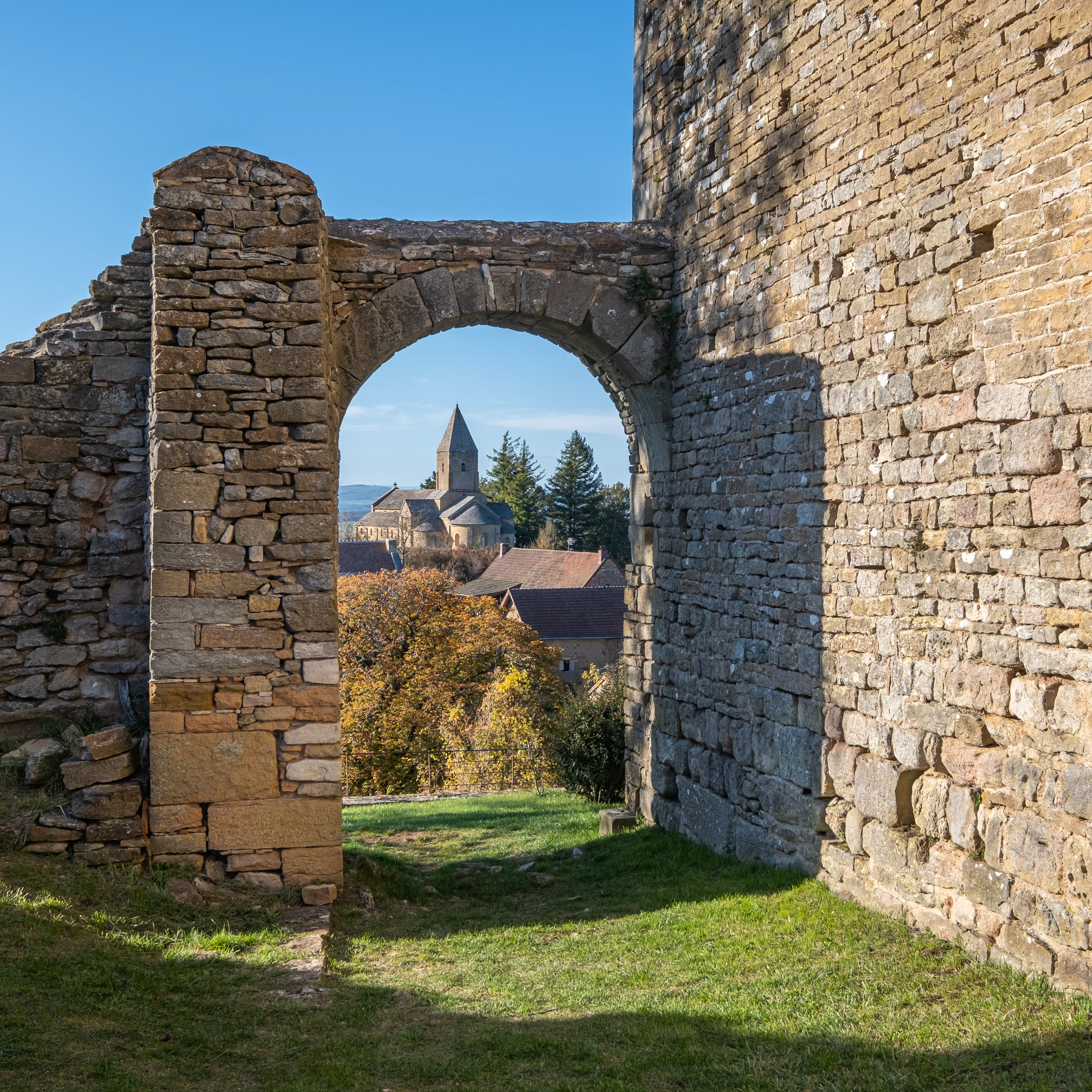 Château de Brancion vu à travers une arche en pierre, avec clocher de l'église romane, bâtiments traditionnels et feuillage automnal, sous ciel bleu clair. Patrimoine architectural et histoire de Saône-et-Loire, Bourgogne. Photographie touristique par Etienne Gatheron.