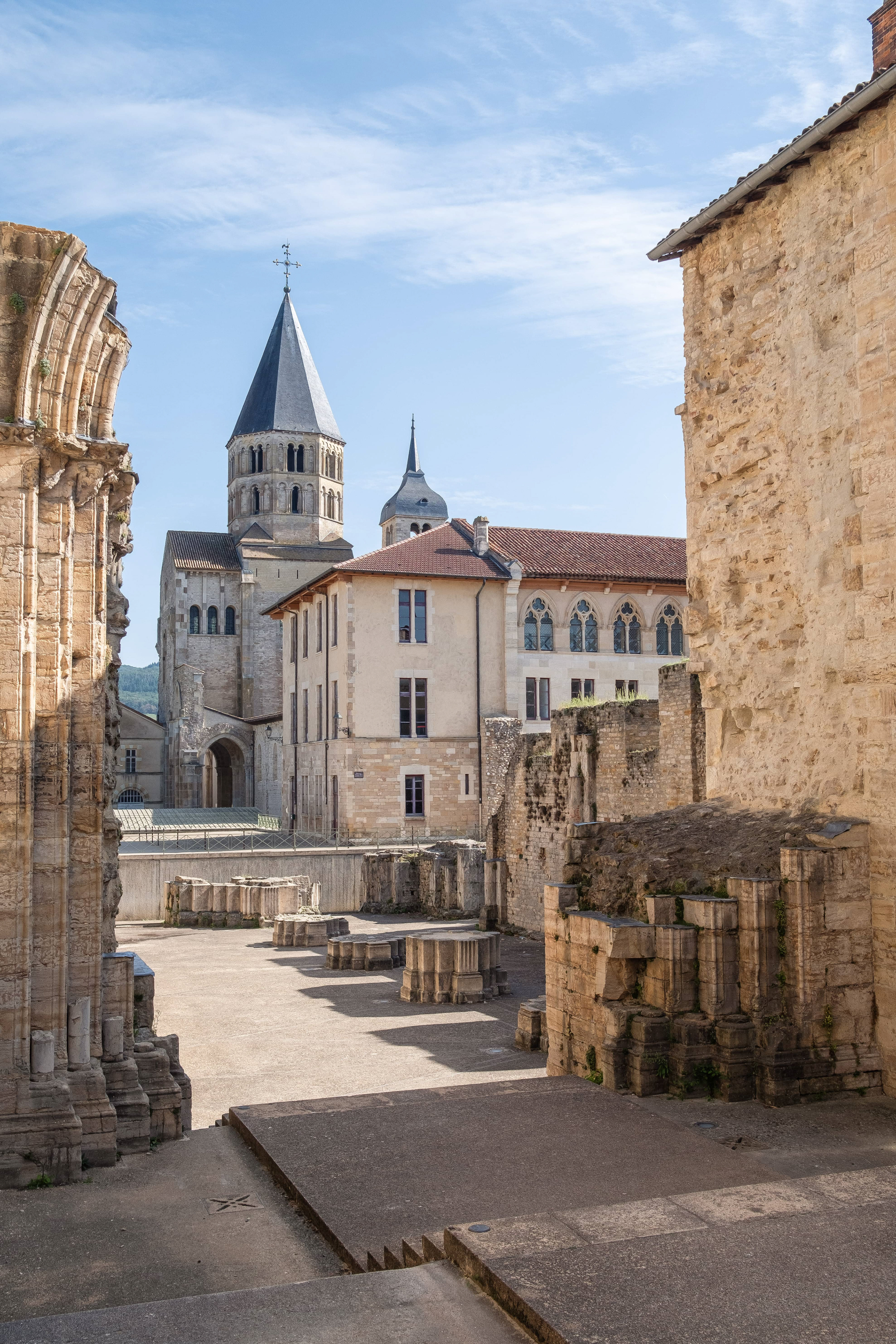 Vue de l'Abbaye de Cluny en Saône-et-Loire, Bourgogne. La photo montre les ruines anciennes de l'abbaye, avec ses colonnes de pierre et ses murs historiques sous un ciel bleu clair. En arrière-plan, le clocher de l'église se dresse fièrement, entouré de bâtiments en pierre bien conservés. Cette scène historique met en valeur l'importance culturelle et religieuse de la région. Photographie touristique réalisée par Etienne Gatheron, photographe institutionnel spécialisé dans les sites historiques et le patrimoine culturel.