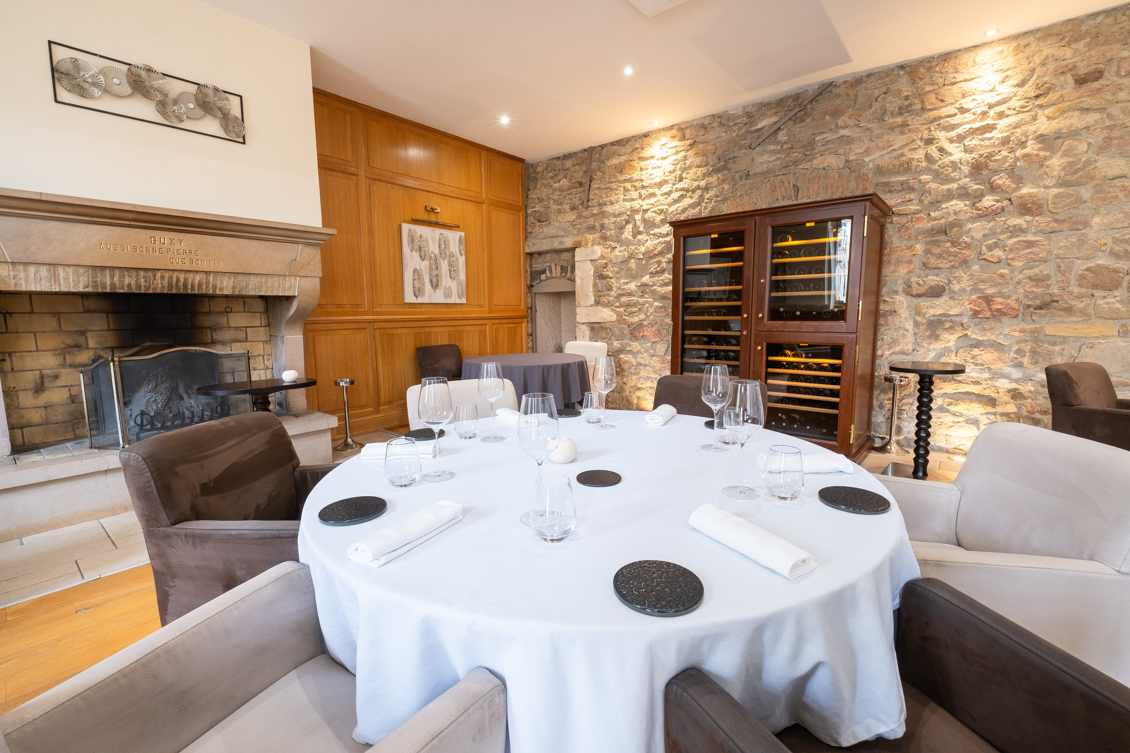 Salle à manger de L'Empreinte à Buxy en Saône-et-Loire, Bourgogne, avec une grande cheminée en pierre, des murs en pierre apparente et en bois, une table ronde élégamment dressée, et une armoire à vin vitrée, créant une ambiance chaleureuse et sophistiquée pour un repas convivial.