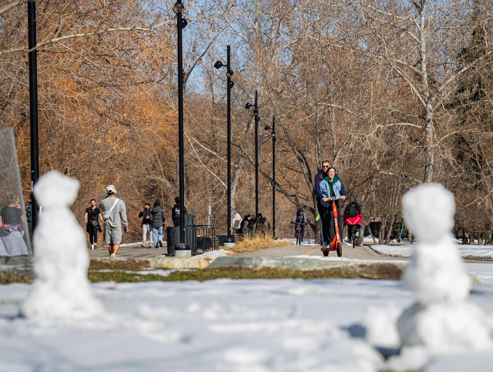 A couple rides an e-scooter on a pathway near the Bow River in Calgary on Monday, April 1, 2024. With the snow sculptures melting away and weather forecasts predicting warmer temperatures this week onwards, Calgarians can expect to see increased activity near the Bow River in the upcoming days, making it a popular destination for recreation. (Photo by Armaan Khanna/The Press)