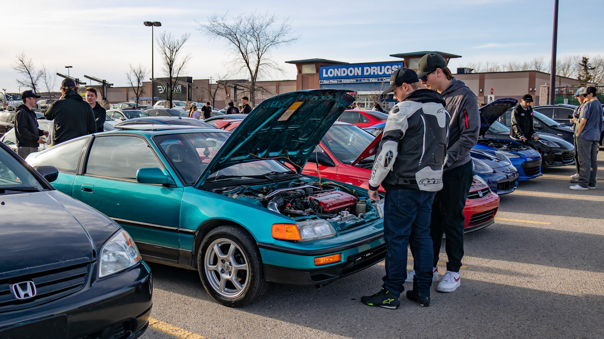 Mike, second to left, the owner of the old modified Honda coupe, chats with passersby gathered to admire modified cars at an unofficial car meet-up in the Royal Oak Shopping Centre parking lot in NW Calgary on Sunday, April 14, 2024. The lot filled up with a variety of modified vehicles displayed by car enthusiasts. The fun ended at sunset when the police showed up. (Photo by Armaan Khanna/The Press)