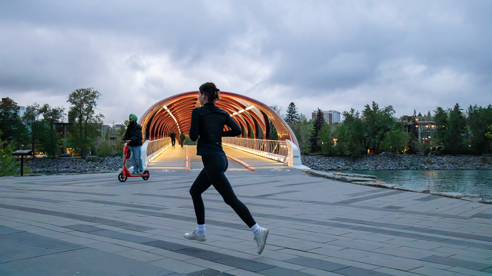 A woman runs in front of the Peace Bridge on a pathway near the Bow River in Calgary on a cloudy and windy Friday evening, May 17, 2024. Popularly known as the Bow River Pathway, it is a very famous place for recreational running, cycling, and walking. The area is peaceful, with many birds chirping. (Photo by Armaan Khanna/SAIT)
