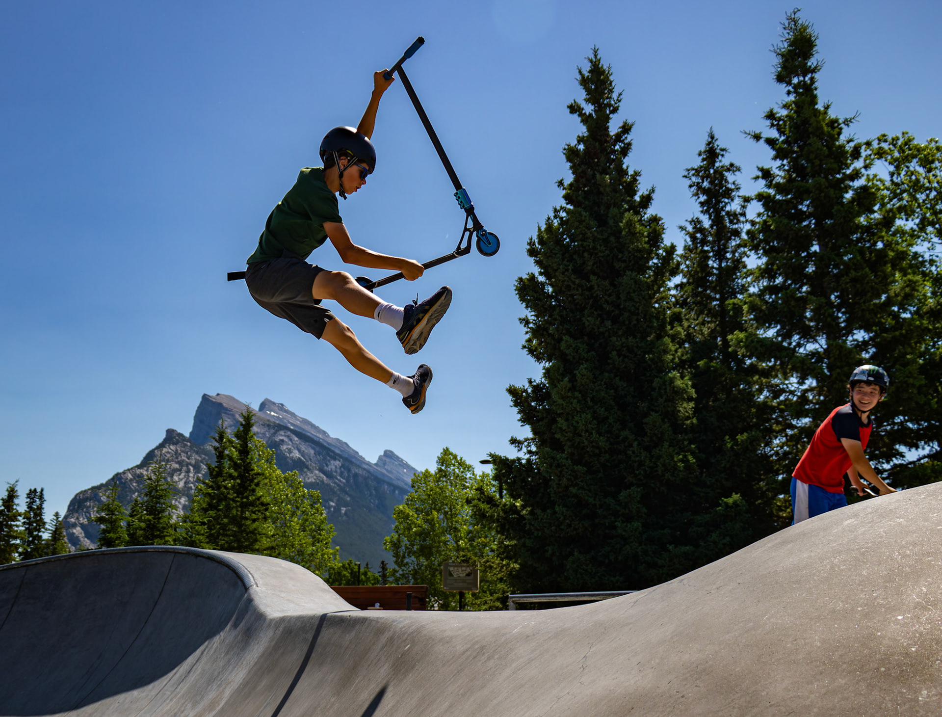 Anders Jhonathan Leif Karlos performs a daring stunt on his scooter, catching air while Liam Duncan watches him at the BanffLIFE Skatepark Community BBQ in the town of Banff, Alberta, on Thursday, July 11, 2024. The event featured soda and hotdogs, bringing the community together for a day of fun and excitement. (ARMAAN KHANNA RMO PHOTO)