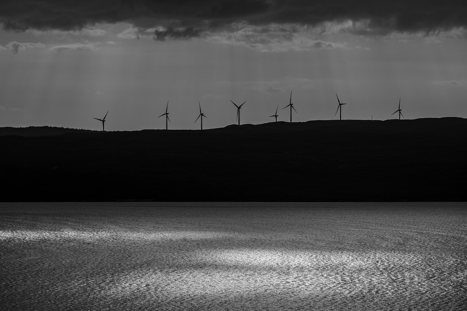 Black and white photograph taken from above Pirnmill on the Isle of Arran, showing wind turbines in silhouette on the mainland with beams of light breaking through clouds and reflecting on the rippling surface of the Kilbrannan Sound.