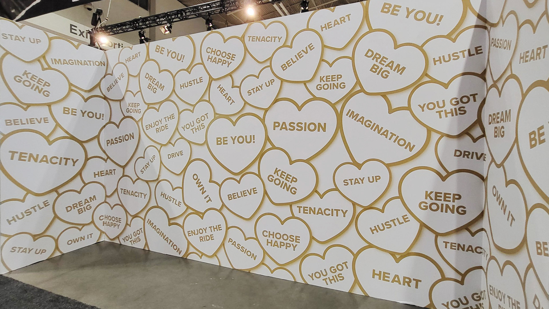 A large white wall in the convention centre that is covered in gold hearts with inspirational phrases in them, such as "Be You!", "Keep Going", "Dream Big", and "You Got This".