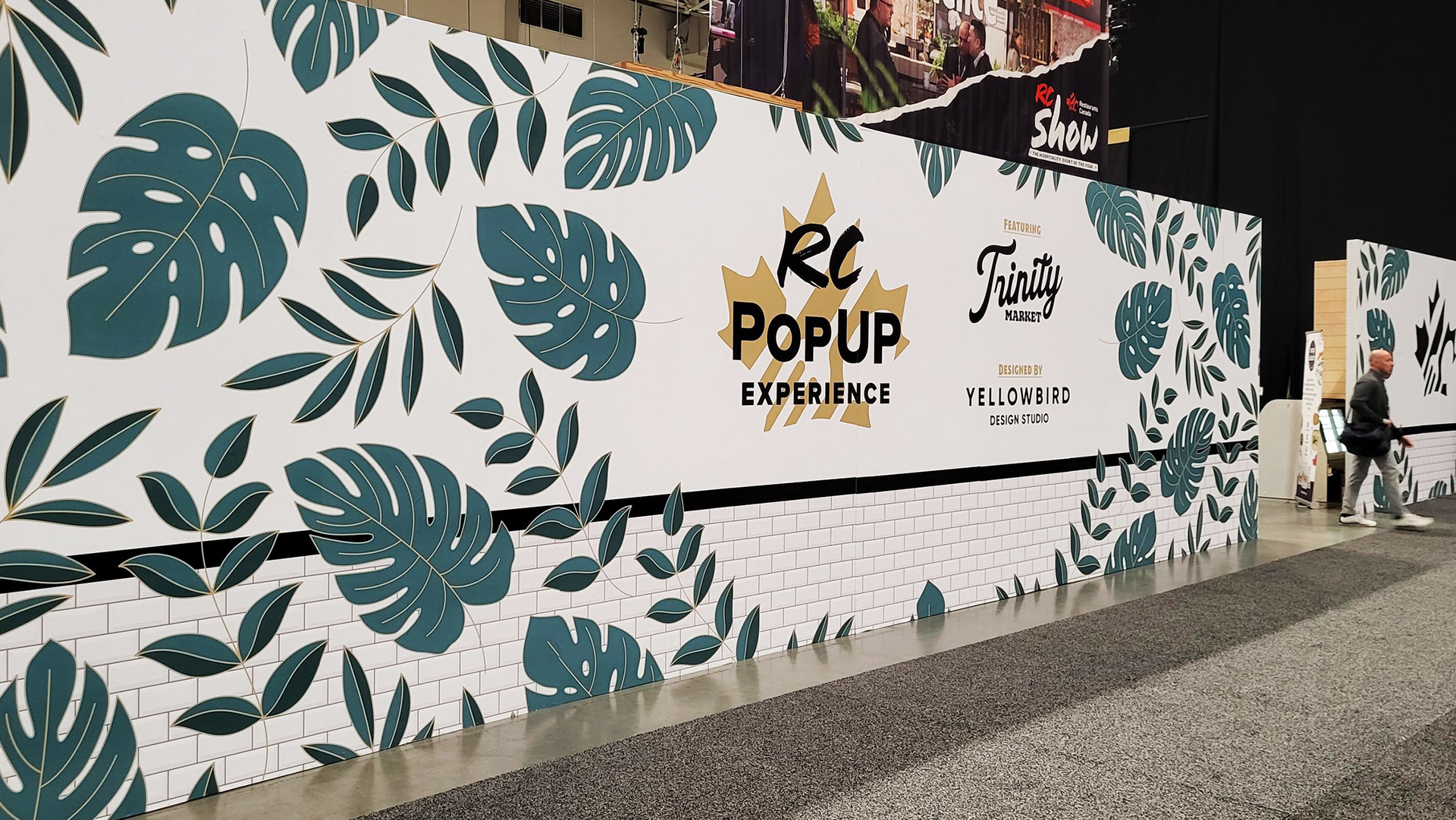 A large white wall with an RC Pop Up Experience logo in the middle, with a stylized tropical leaf pattern design surrounding it.