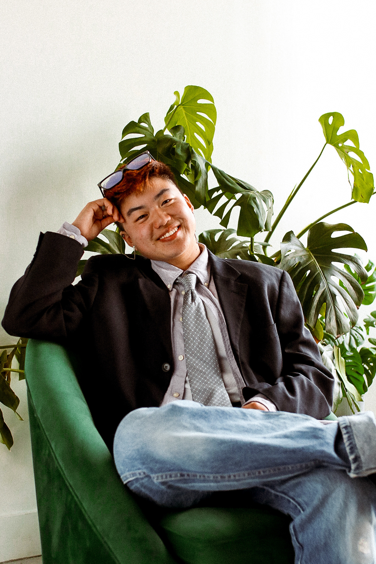 A young Asian-American person sits in a dark green chair in front of a white wall. Behind them are bright green potted plants.