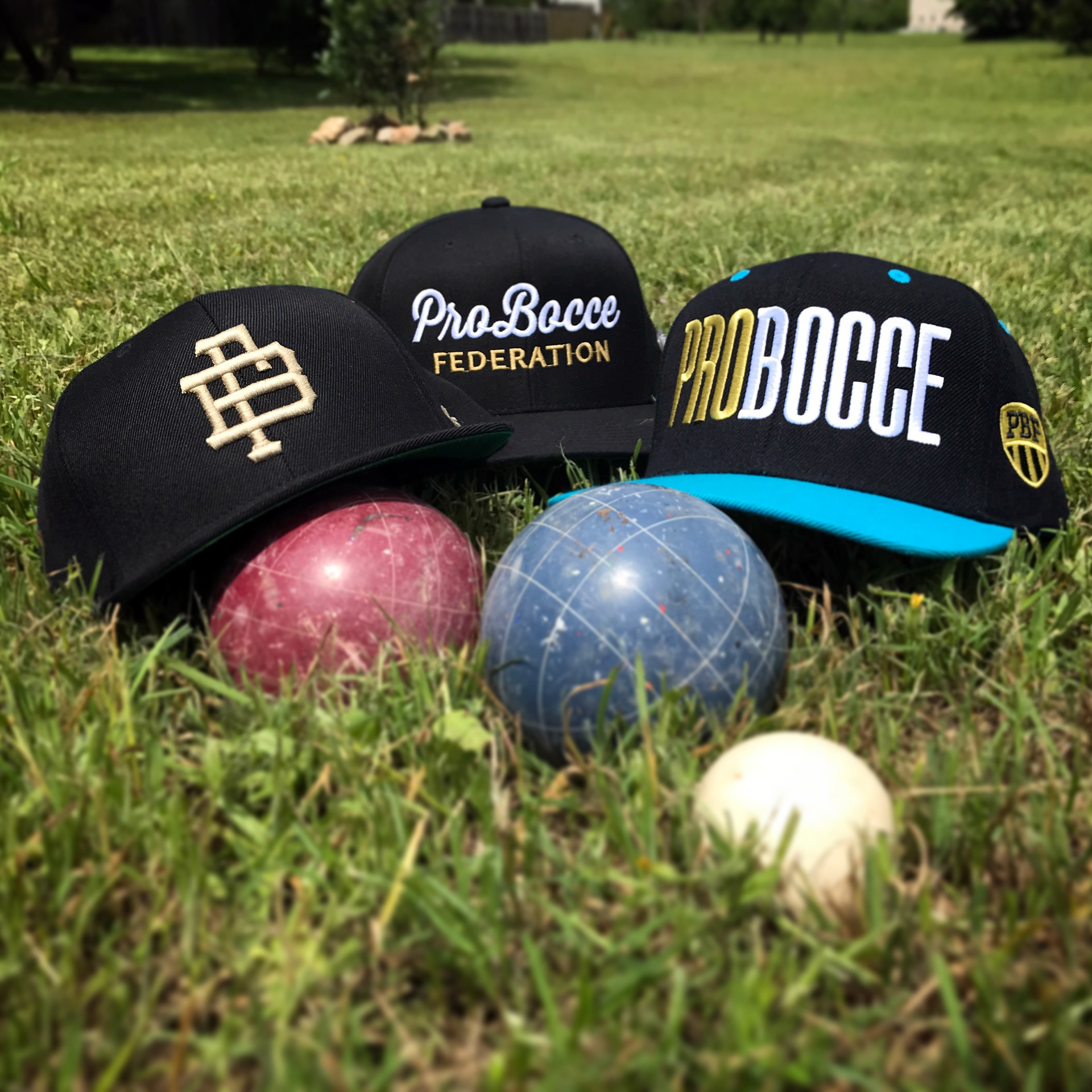 3 bocce-themed snapback caps sit behind 2 bocce balls on a luch green field