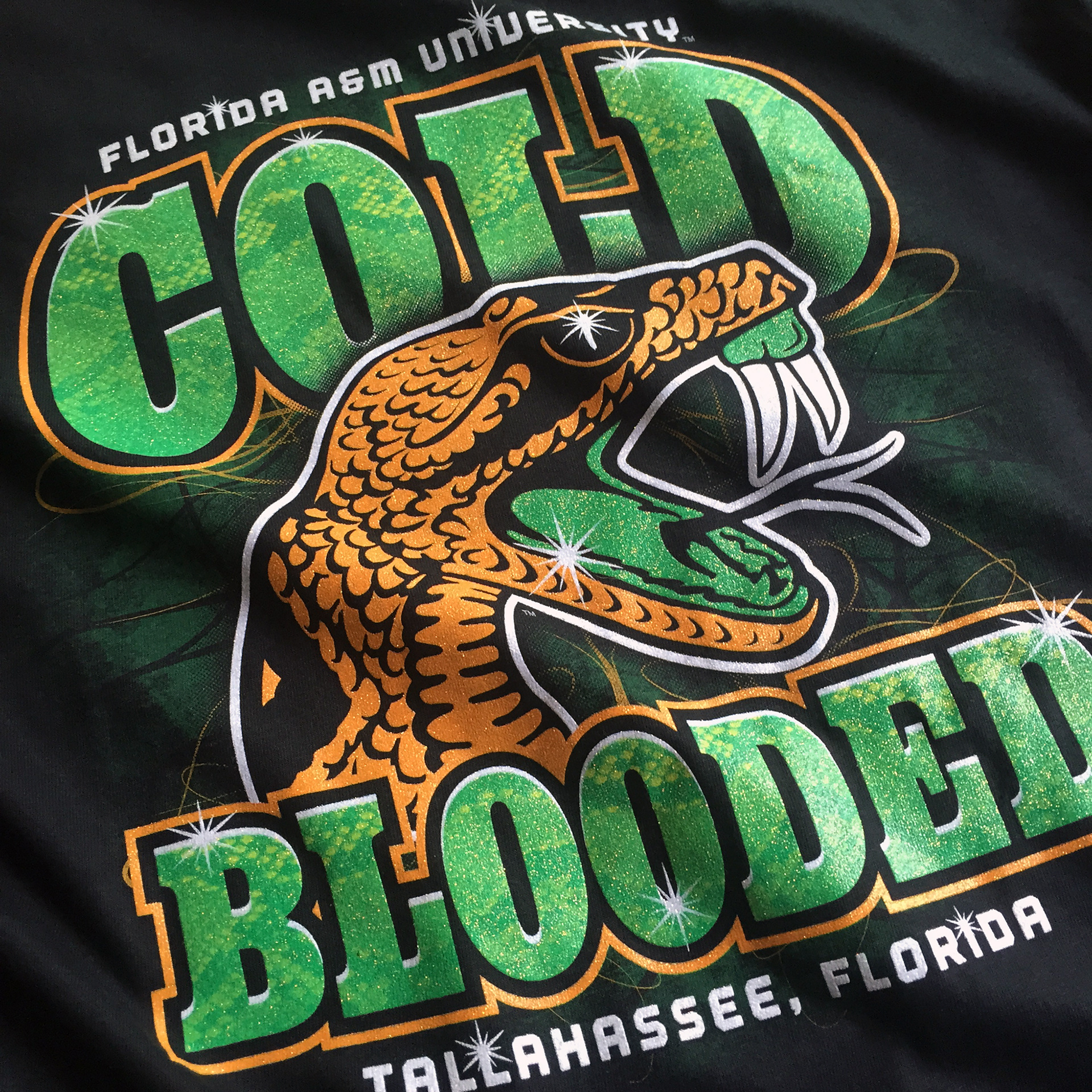 A detailed photo of printed, licensed collegiate merchandise: "COLD BLOODED" coupled with the Florida A&M Rattlers athletic logo; with a snakeskin pattern and crystalina ink.