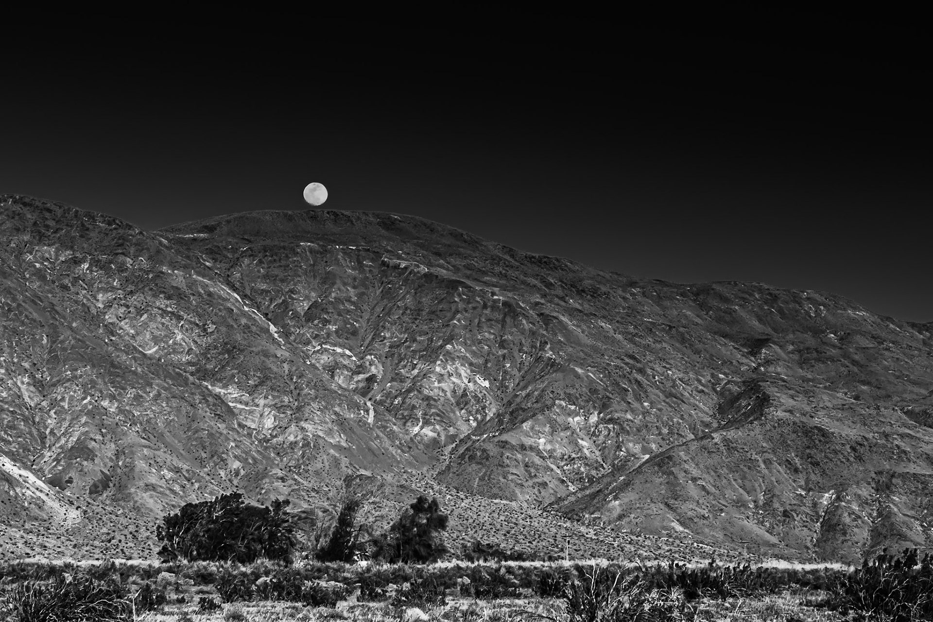 Evening Moonrise Black-and-White: Viewed across the creosote-bush studded, alluvial plain near the entrance to Coyote Canyon in the Anza-Borrego Desert.  A house nestled in a copse of willows sites at the far side of the plain at the foot of the mountain.