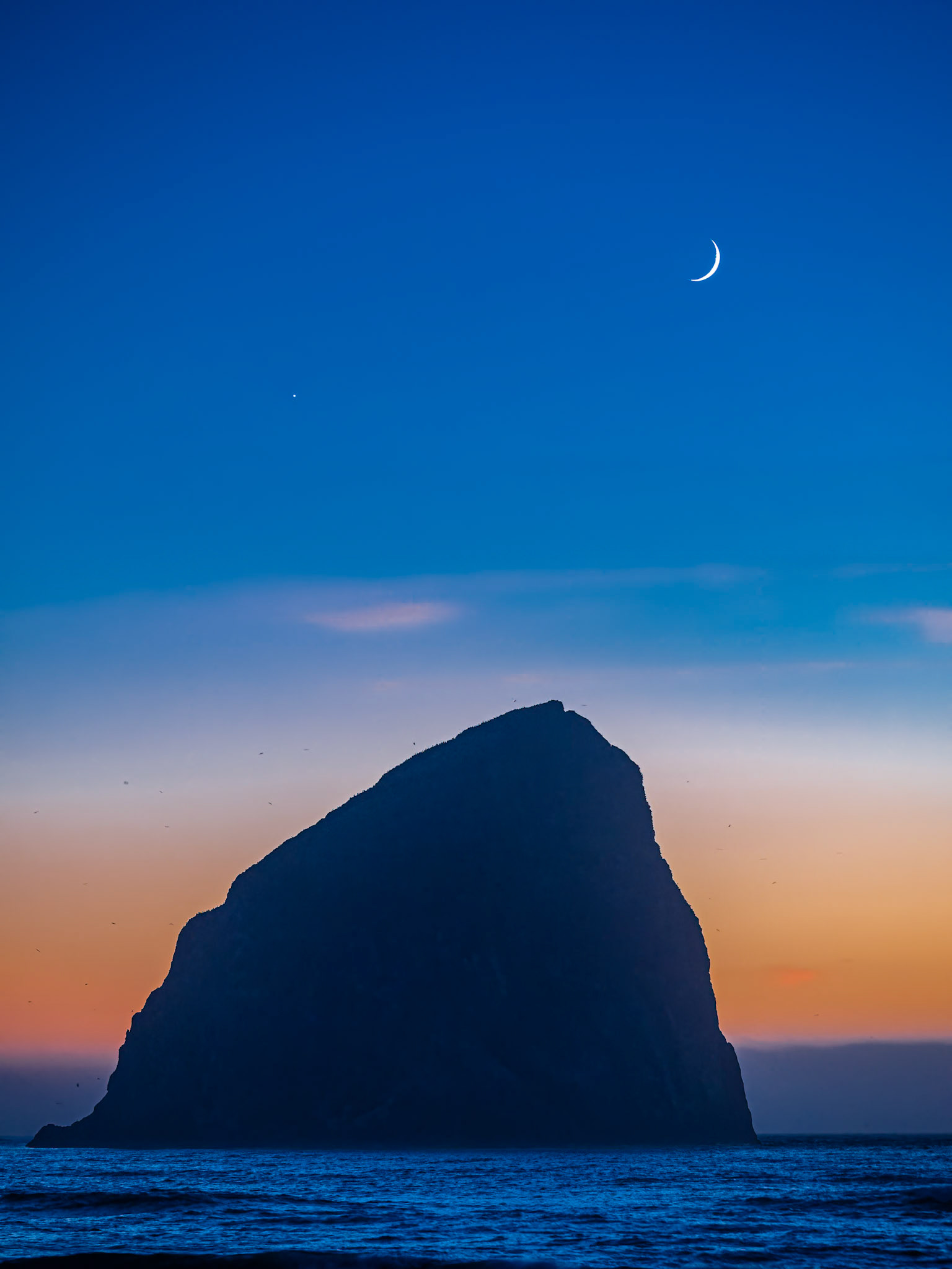 Chief Kiwanda Rock Under a Crescent Moon - Portrait: The crescent moon and Venus setting behind Chief Kiwanda Rock in the blue hour after sunset in Pacific City Oregon.